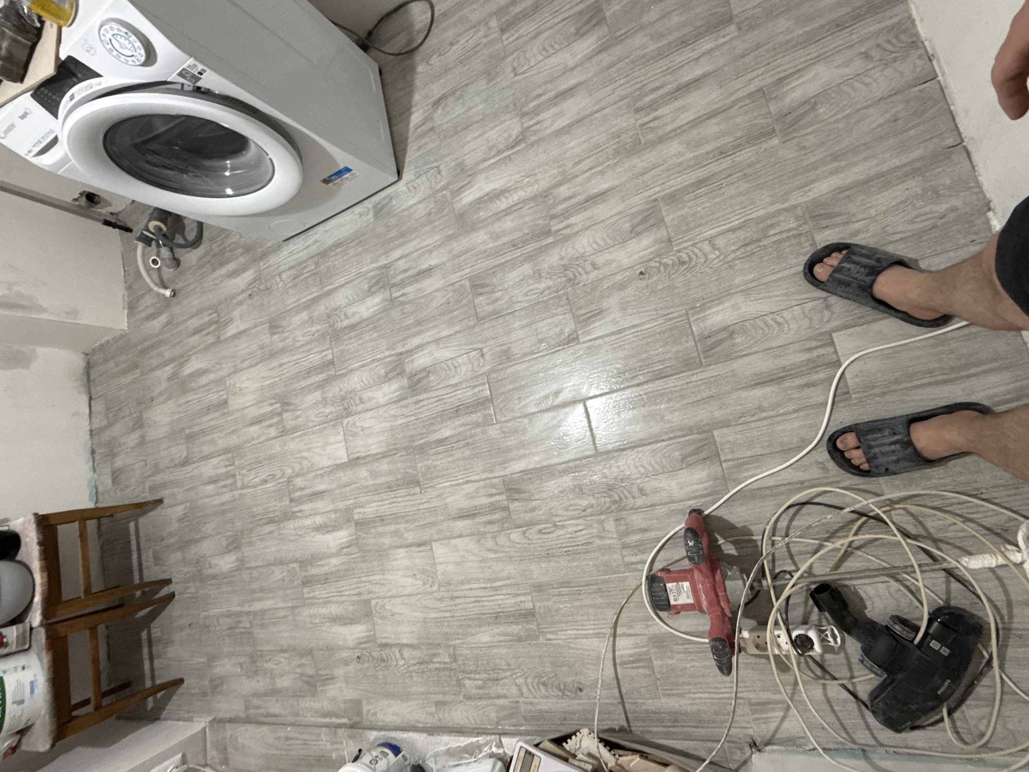 The scene is an indoor space, likely a utility room or laundry area, featuring a white washing machine in the upper left corner. The floor is covered with light gray, wood-patterned tiles. A small, wooden shelving unit is visible along the left wall.
To the right of the center are two feet wearing dark-colored sandals. Dispersed on the floor near the feet are various power tools, including a red detail sander and a black hand vacuum, along with a white multi-socket power strip and a tangle of electrical cords.
The location is not identifiable as Stara Zagora, Bulgaria, from the image.