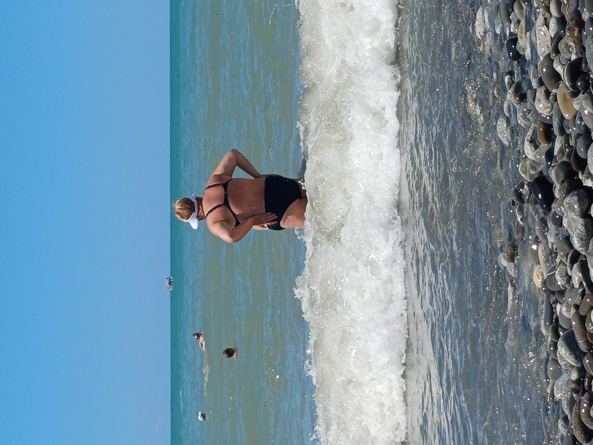 An eye-level, medium shot captures a woman in a black bikini and white visor standing in the shallow ocean water. She is facing away from the camera, with her left hand on her hip and her right arm bent with her hand on her hip as well. She is standing in a moderate wave breaking around her knees. In the background, the vast turquoise ocean extends to the horizon, meeting a clear blue sky. Several other people are visible further out in the water, some swimming, and a small boat or raft is present on the horizon. The foreground is dominated by a pebbly beach, with smooth, rounded stones of various sizes and colors. The overall scene depicts a summer day at the beach.