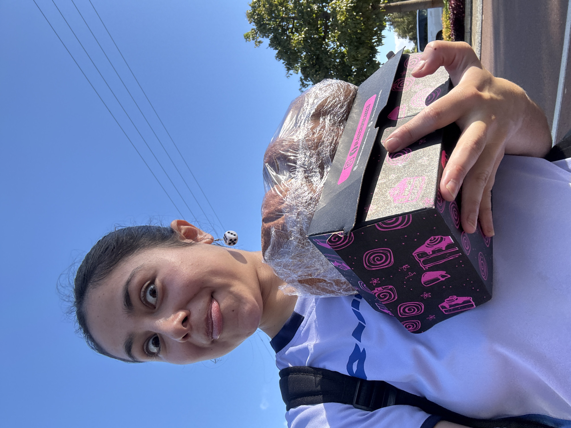 A person, likely female, is holding a box of baked goods wrapped in plastic in an outdoor setting. The box is black with pink dessert-themed designs. The person has dark hair pulled back, is wearing a white shirt with blue details, and has dice-shaped earrings. A backpack strap is visible. Background features include blue sky, power lines, a tree, and a glimpse of a building or vehicle. There is no direct indication of Bassendean, Australia in the picture.