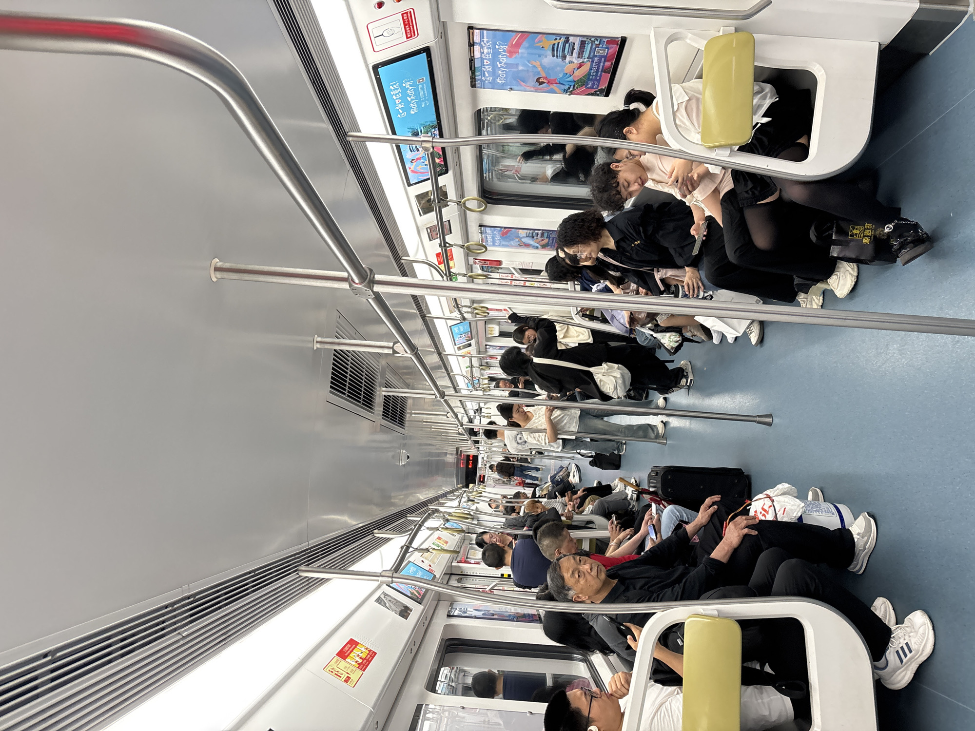 The interior of a subway car, possibly in the Jiulongpo District, China, is shown with rows of seated passengers along each side. Handrails run along the ceiling and down to the seats. Displays are visible above the seats, showing text in Chinese and images. The color scheme is largely white and grey, with a blue floor. People are sitting with belongings next to them. Some are looking at their phones.