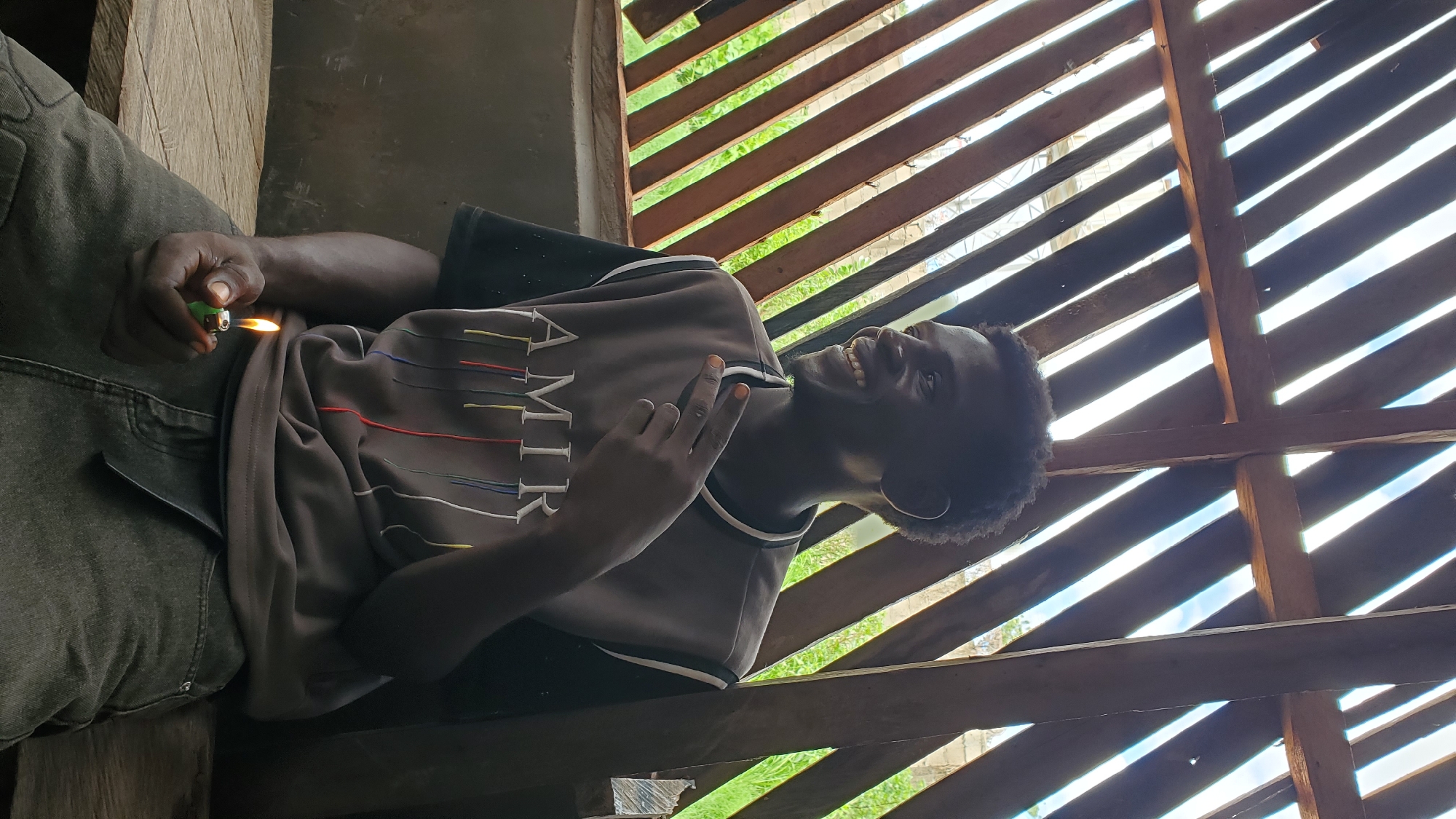 A young Black man is seated on a wooden bench under a slatted wooden roof. He is smiling and looking to his right. His right hand is raised, and he is holding a lit green lighter between his thumb and index finger. He is wearing a dark t-shirt with a grey sleeveless top over it, which has "AMIRI" printed on the front in white letters with colorful vertical lines below. He is also wearing dark-colored pants. The background shows the sky and some greenery visible through the slats of the roof. The scene appears to be outdoors, possibly in a casual outdoor seating area in Jalingo, Nigeria.