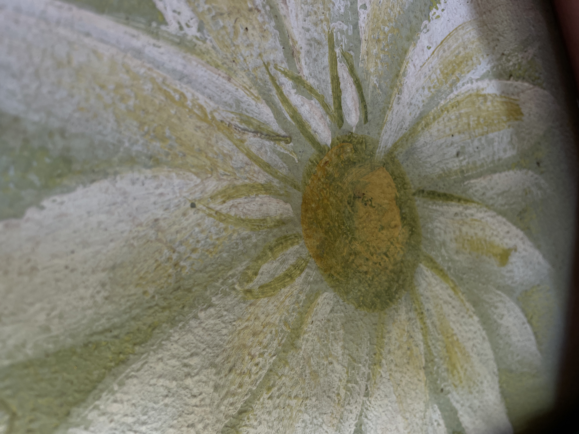 A close-up, macro photograph depicts a painted white daisy flower with a yellow and green center. The flower's petals are textured with visible brushstrokes and a slightly rough surface. The central disk of the flower exhibits a mottled yellow and olive green hue. The background is a soft, muted green with hints of white, suggesting a painted surface. The lighting is soft and diffused, casting subtle shadows that highlight the texture of the paint. The image is a detail of a larger artwork, with no specific city or location information discernible.