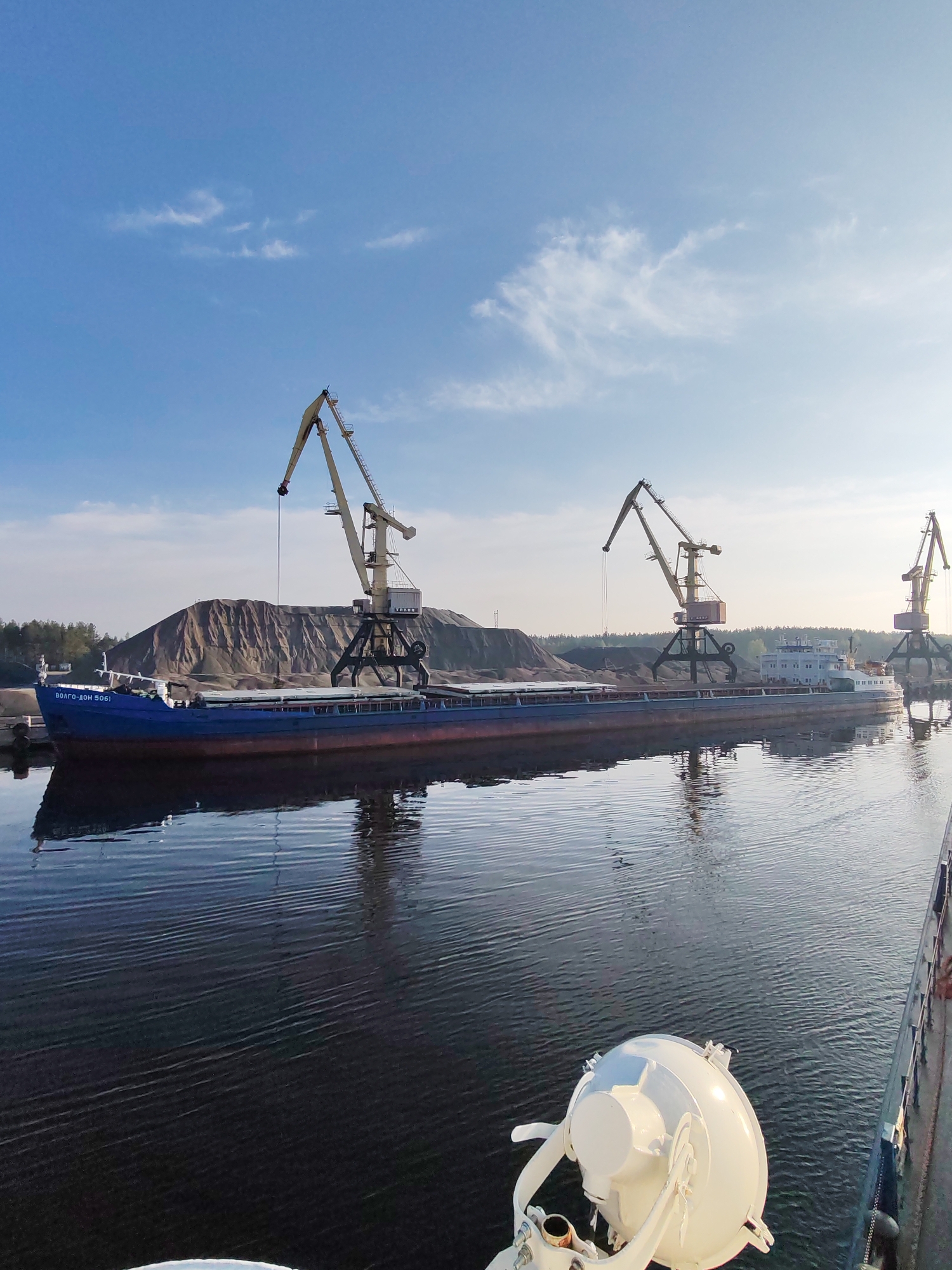 The image shows a blue and maroon cargo ship named "Volgo-Don 5061" docked in a port, likely in Shalsky, Russia. There are three large, yellow harbor cranes positioned along the ship's length. Behind the ship and cranes, there is a large pile of grayish-brown material, possibly sand or gravel. The water is dark and still, reflecting the sky and objects along the shoreline. The sky is blue with scattered white clouds. A white, spherical light fixture is visible in the foreground on the right side of the image, suggesting the viewpoint is from another vessel. The overall scene suggests a cargo loading or unloading operation in a port environment.