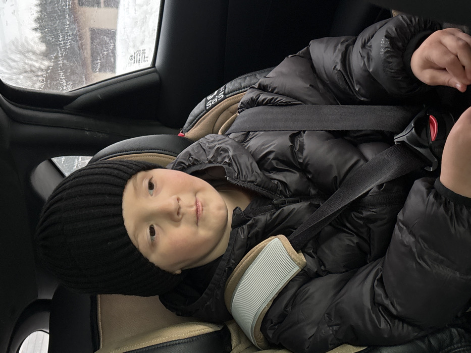 A young child, approximately 4-7 years old, is seated in a car seat within a vehicle in Pavlovsky, Russia. The child, with fair skin and light brown hair partially visible, wears a black ribbed knit beanie and a black puffer jacket over a dark shirt. The car seat is beige and black, featuring a grey and white striped shoulder pad on the left strap. A black seatbelt is securely buckled across the child's chest and lap, with a red release mechanism visible. The child's gaze is directed slightly to the viewer's right, towards the car window. Through the window, a snowy outdoor environment is visible, featuring snow-covered ground and bare trees or shrubs, with a vague brown structure in the background, indicative of winter conditions. The vehicle's interior appears dark-colored.