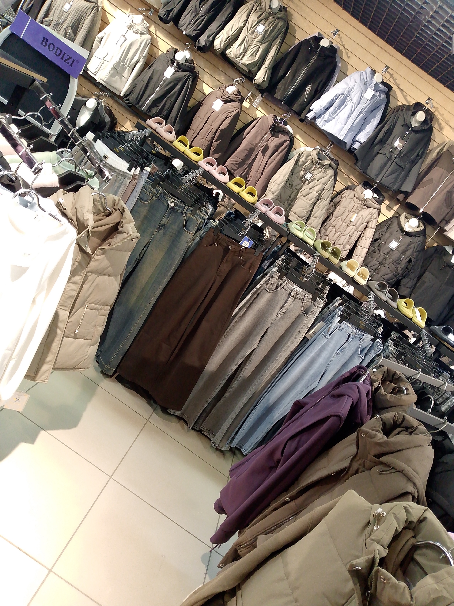 This is a high-angle shot of a retail clothing store. The store appears to be located in Magadan, Russia, based on the purple sign that reads "BODIZI". The shelves and racks are filled with various items of clothing, including puffer jackets, denim jeans, and what appear to be dress pants. The clothing is neatly displayed and organized by color and type. The store has a clean and modern aesthetic, with light-colored tiled flooring and wooden paneled walls. There are no people visible in the image. The lighting is bright and even, indicating artificial lighting.