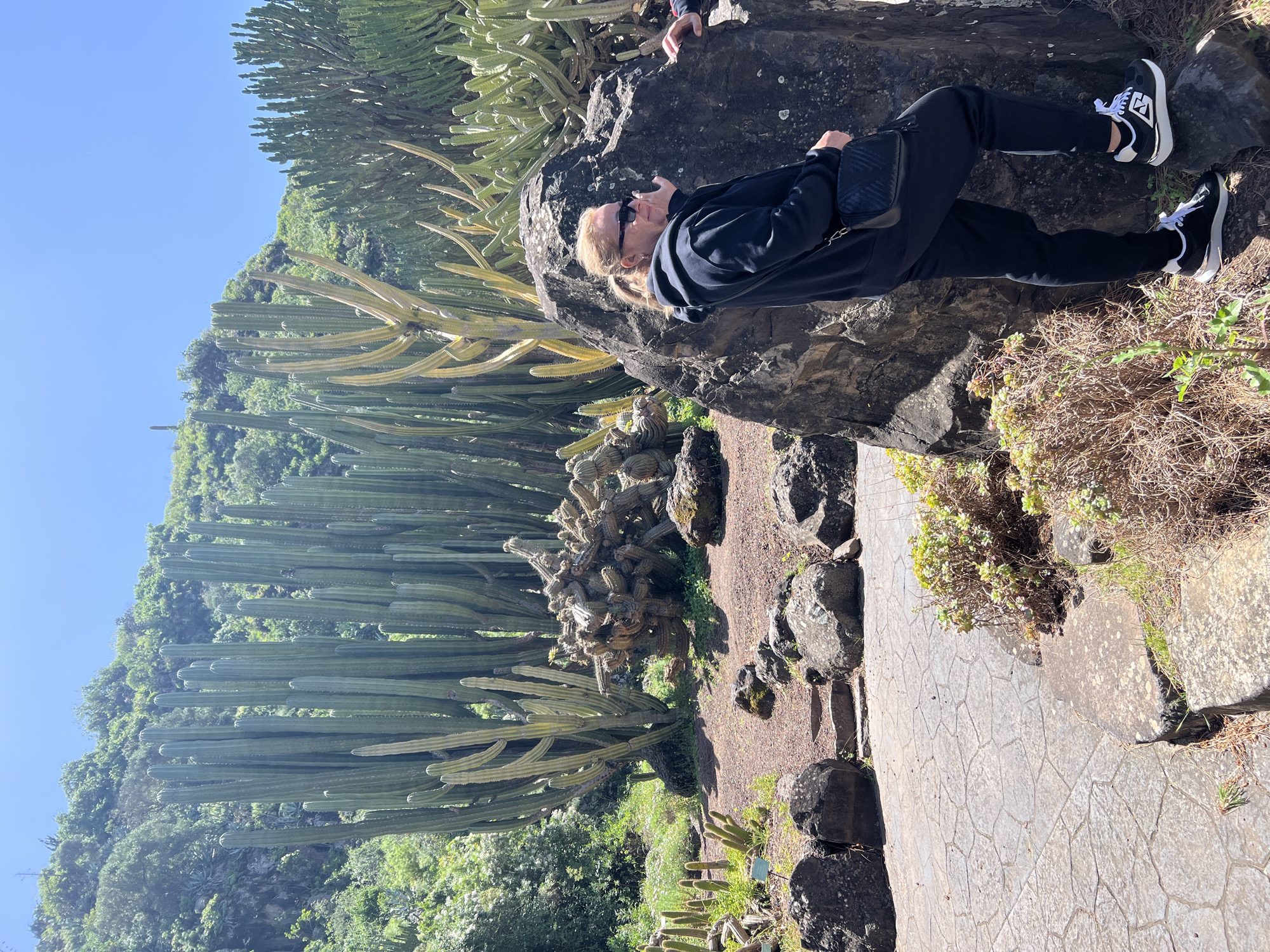 A middle-aged woman lies on a large rock formation in a botanical garden in Las Palmas, Spain. She wears sunglasses, a black jacket, black trousers, and black and white sneakers. A black shoulder bag rests near her waist. A person's hand is visible on the upper edge of the rock. The path is paved with grey stones and is edged with dark rocks. Tall cacti stand beside the path and climb the hillside behind. A cloudless blue sky is visible.