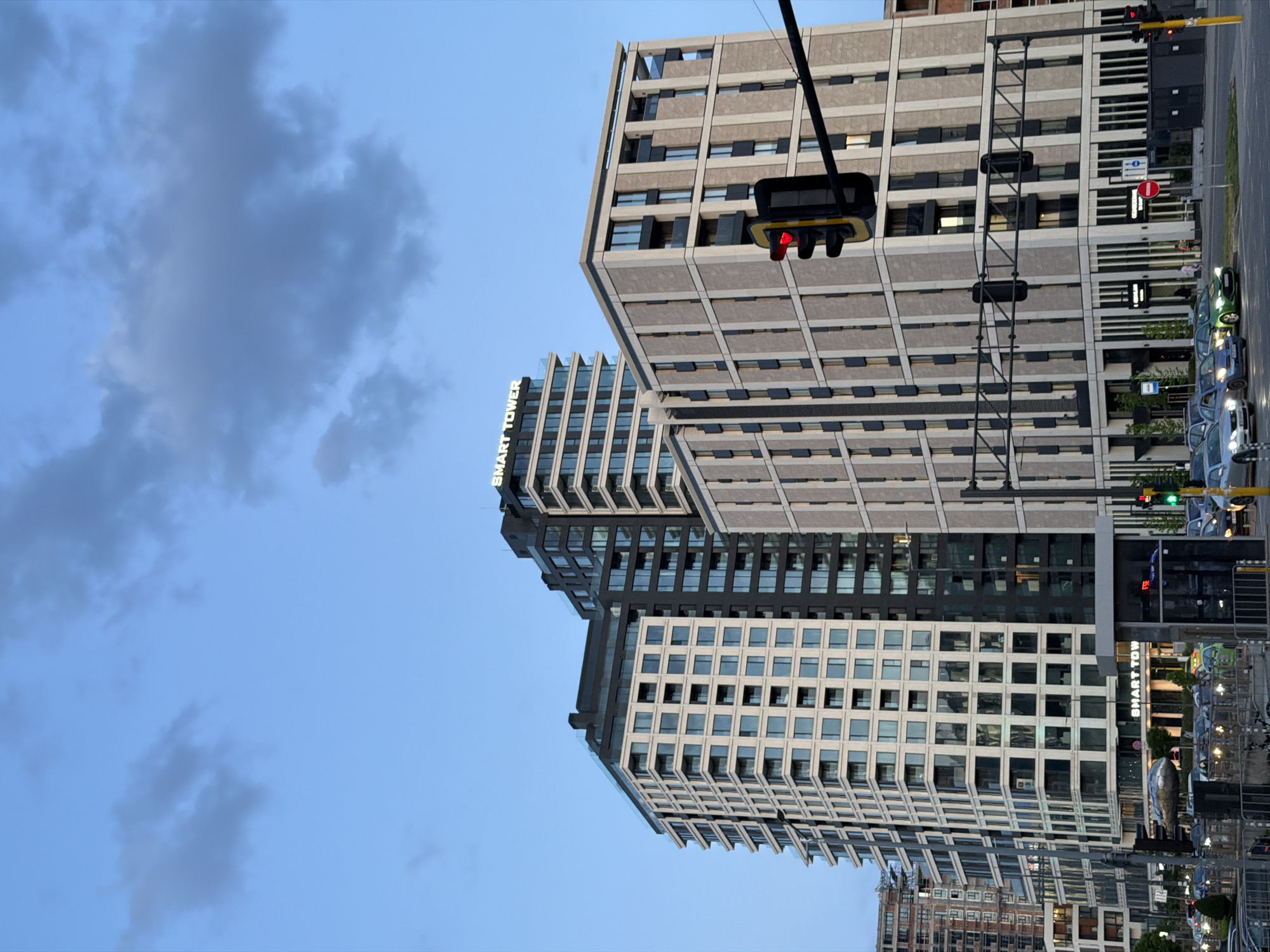 The image depicts two modern high-rise buildings, with a traffic light in the foreground displaying a red signal. The buildings have a grid-like design with alternating dark and light-colored panels. One building displays the name "SMART TOWER" vertically. A city street with parked cars runs alongside the building. The sky is blue with some clouds. The location is likely Sofia, Bulgaria, due to the text "SMART TOWER" and the architectural style.