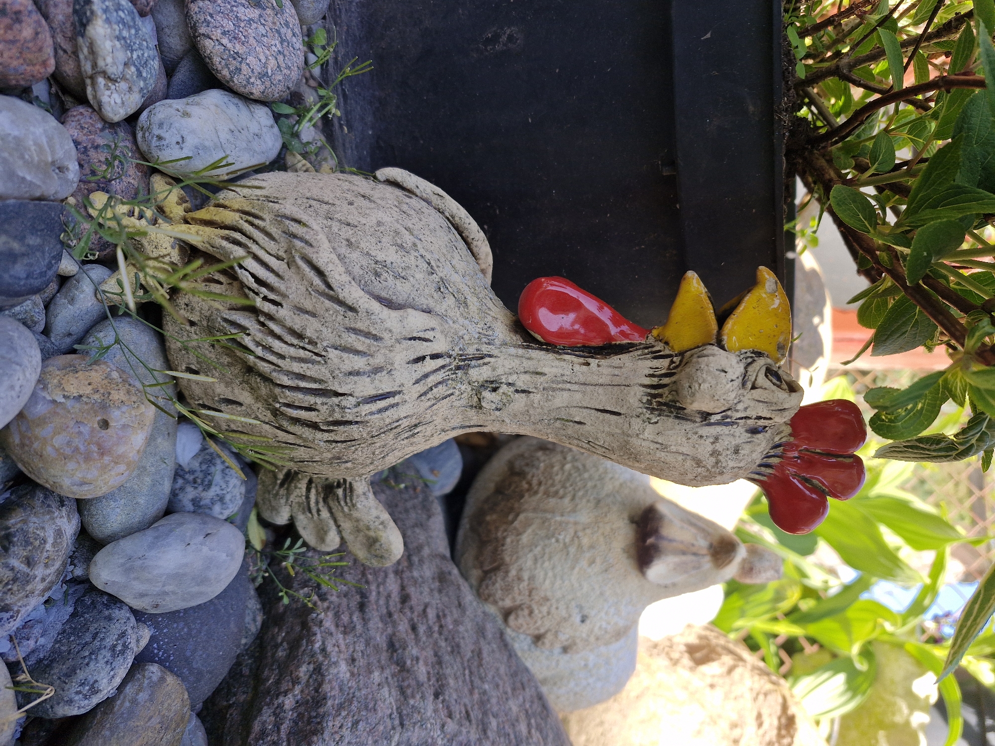 The image shows two ceramic figurines, a rooster and a small sheep-like animal, placed amongst rocks and plants. The rooster is primarily gray with red and yellow accents on its head. It is positioned near a black container and some greenery. The smaller animal is light beige and situated near the lower edge of the frame, adjacent to a large rock. The setting appears to be an outdoor garden or similar space.