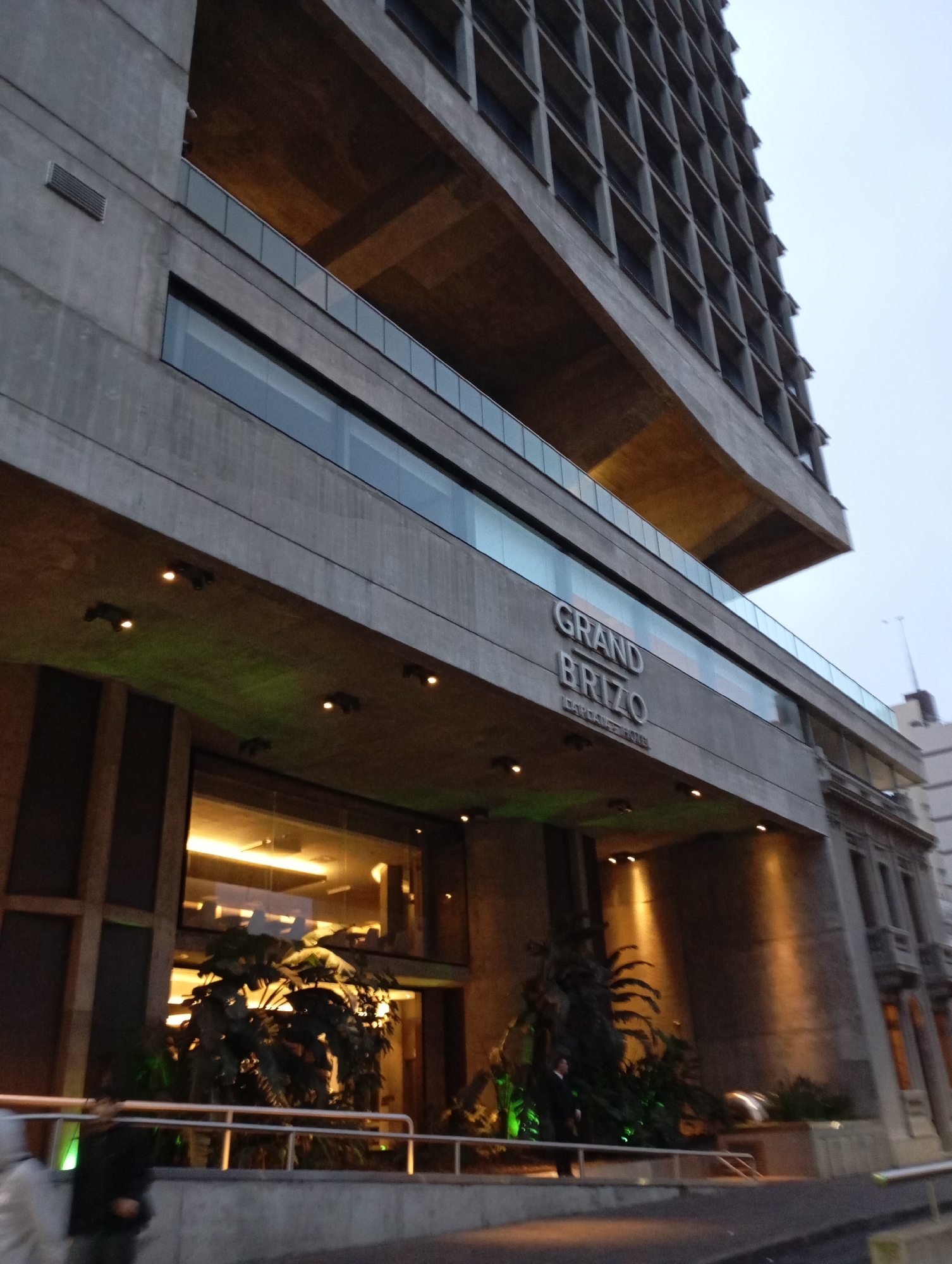 The image shows a low-angle view of a Brutalist-style hotel, the Grand Brizo, located in the city of La Plata, Argentina. The building is characterized by its rough concrete facade, cantilevered floors, and horizontal bands of windows. The entrance area is visible, featuring large glass windows, internal lighting, and tropical plants. Several small, dark lights are mounted on the underside of the overhanging structure, casting a greenish glow. A railing runs along the front of the building. In the foreground, two blurry figures can be seen, one wearing a hooded jacket and another in a dark suit, appearing to be employees or guests. The sky above is overcast.