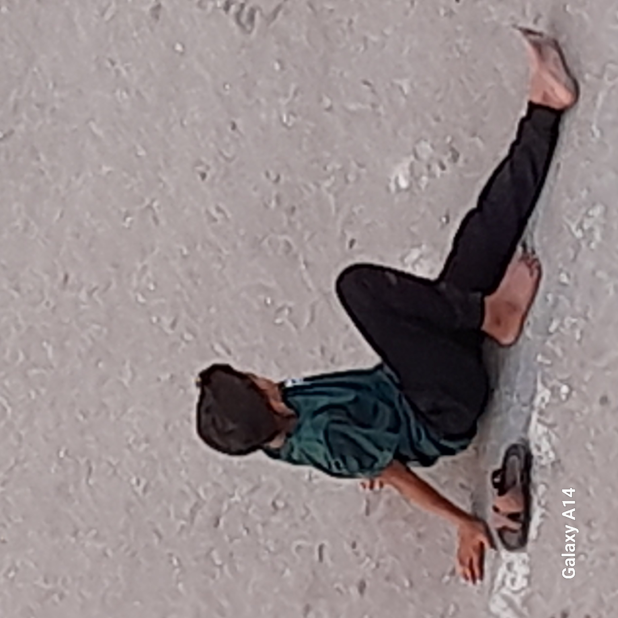 The image shows a person lying on a sandy surface, likely a beach or a dry, outdoor area. The person is wearing black pants and a dark green shirt. One foot is extended, with toes visible, and the other foot is partially obscured by the body. A sandal is lying on the sand near the person's feet. The background is a textured, sandy surface with some small pebbles or debris. The overall lighting suggests an outdoor environment during the day. The text "Galaxy A14" is visible on the right side of the image, indicating the device used for capture. There are no visible structures or clear indicators of a specific urban location like Khan Yunis.