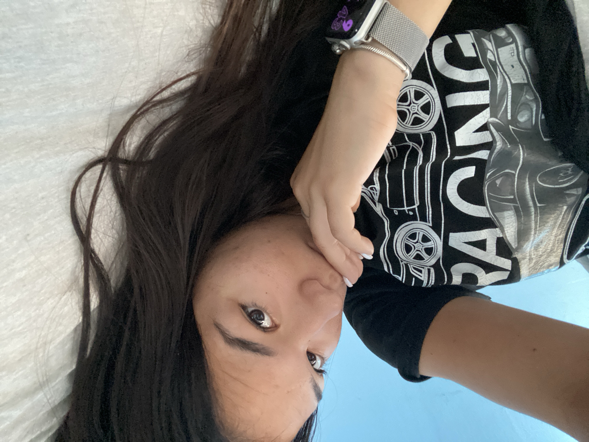 A young woman with dark hair lies on her side on a white surface, looking towards the camera. She is wearing a black t-shirt with a graphic print of a car and the word "RACING" in white letters. Her right hand is raised to her mouth, and she is wearing a silver smartwatch on her left wrist. The background is a plain blue surface. The image appears to be a selfie taken indoors. There are no discernible details that would indicate the location in the city of Hirevo, Bulgaria.
