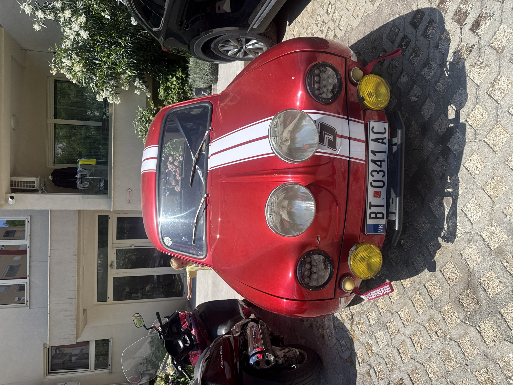 In Boreti, Montenegro, a bright red vintage car with white racing stripes on the hood is parked on a patterned paver surface. The car has large round headlights, one with a circular grille, and two smaller yellow fog lights. A license plate displaying "MK BT 0341 AC" is visible. A red "REMOVE BEFORE FLIGHT" tag hangs from one of the fog lights. To the left of the car, a portion of a large, dark red motorcycle with a prominent windshield is visible. In the background, a modern building with windows and a balcony is present, along with a blooming white-flowered bush. A child with blonde hair, wearing a yellow shirt, stands near the entrance of the building, with their left arm extended towards the car.