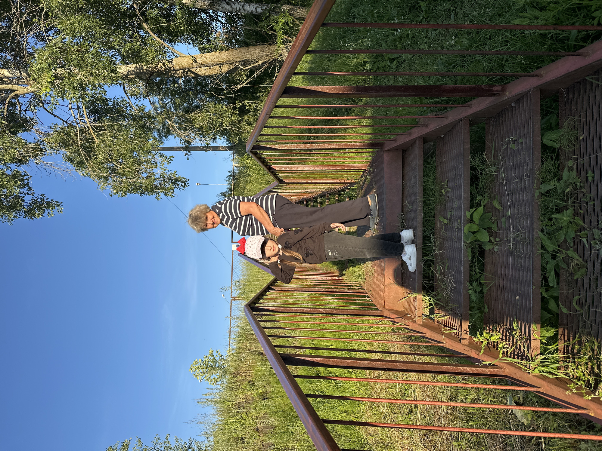 A girl and a woman stand on a metal staircase outdoors. The staircase has a rusty brown railing and is surrounded by green grass and trees. The sky is clear and blue. The girl is wearing a black sweatshirt, dark jeans, and white sneakers. She has a pink and white baseball cap on. The woman is wearing a striped t-shirt and dark pants. The scene appears to be in a park or natural area.