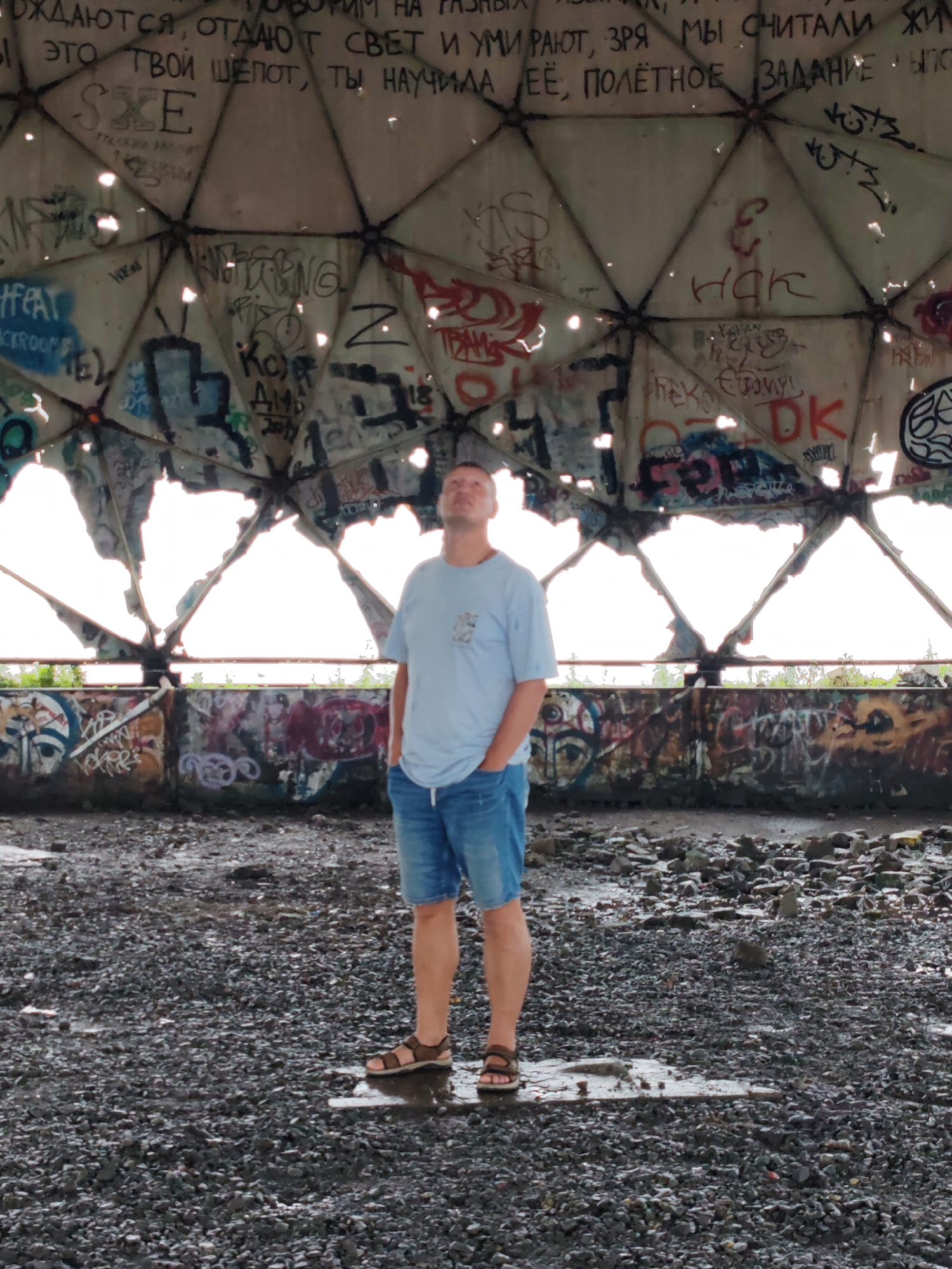 A full shot shows a man standing in the center of a dilapidated geodesic dome structure covered in graffiti. The man is wearing a light blue t-shirt and denim shorts, with his hands in his pockets. He is looking up towards the damaged ceiling of the dome. The floor is covered in gravel and debris, with puddles of water reflecting the light. The dome's structure is visible through large holes in the ceiling, revealing a bright sky. The surrounding walls are also covered in graffiti. This structure is located on Cape Churkin in Vladivostok, Russia, an area known for its abandoned industrial facilities and scenic views.