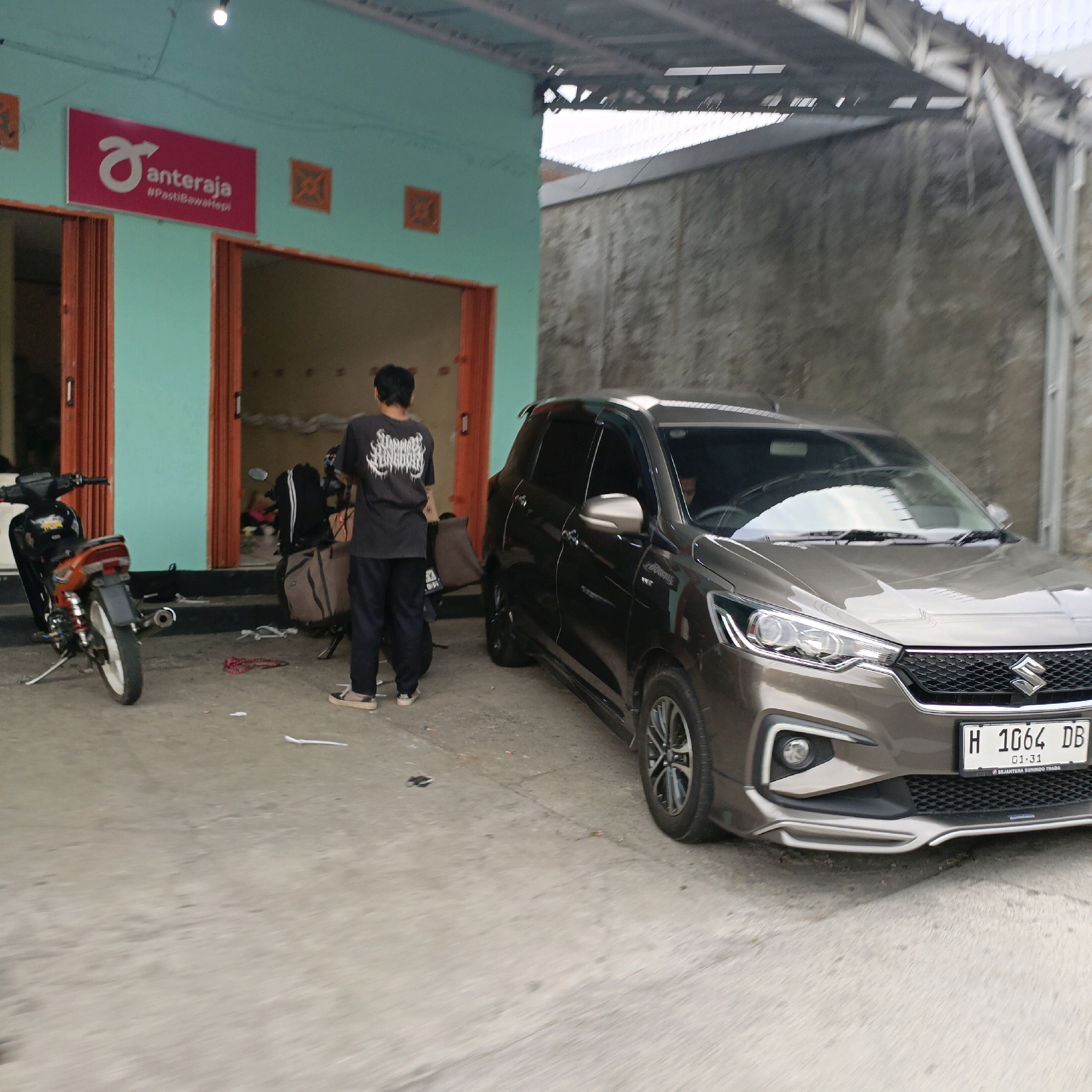 The image depicts an outdoor scene in Salatiga, Indonesia, in front of a teal-colored building with orange-trimmed doorways. A brown Suzuki Ertiga car is parked to the right. To the left, a motorcycle with orange accents is visible. A person wearing a dark t-shirt with white lettering and dark pants stands between the motorcycle and the car, facing away from the camera and towards the building. There are also several large bags leaning against a scooter or another motorcycle behind the standing person. A sign on the building reads "anteraja" with a hashtag "#PastiBawaHebat". The ground is paved, and a gray concrete wall is visible in the background.