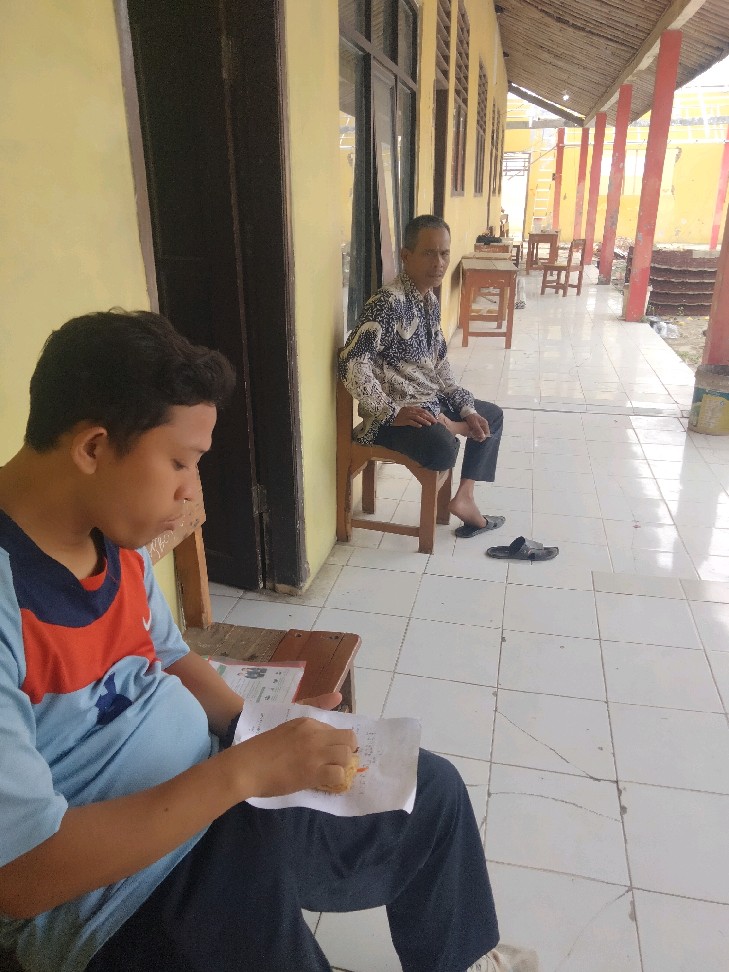 Two individuals are present in an outdoor, covered walkway adjacent to a yellow building. The person in the foreground, seated on a wooden bench, is a young male wearing a light blue t-shirt with red and dark blue accents. He is looking down at a piece of paper in his hands, which appears to have writing on it, and is holding a small, light-colored object, possibly food. The person in the background, seated on a wooden chair, is an older male wearing a patterned long-sleeved shirt and dark pants. He is facing the camera with a neutral expression. The walkway has a tiled floor and is supported by red pillars. Wooden tables and chairs are visible further down the walkway. The general environment suggests a school or community setting in Suka Sari, Indonesia.
