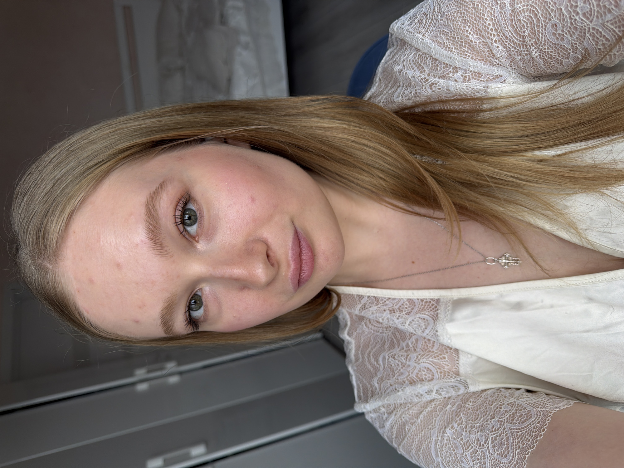 A close-up, portrait-style photograph shows a young woman with light blonde hair, green eyes, and fair skin. She is wearing a white, lace-trimmed garment, and a silver necklace with a pendant is visible. The background includes a gray surface and a white, soft texture. There is no indication of the location being in Rossiyskiy, Russia.