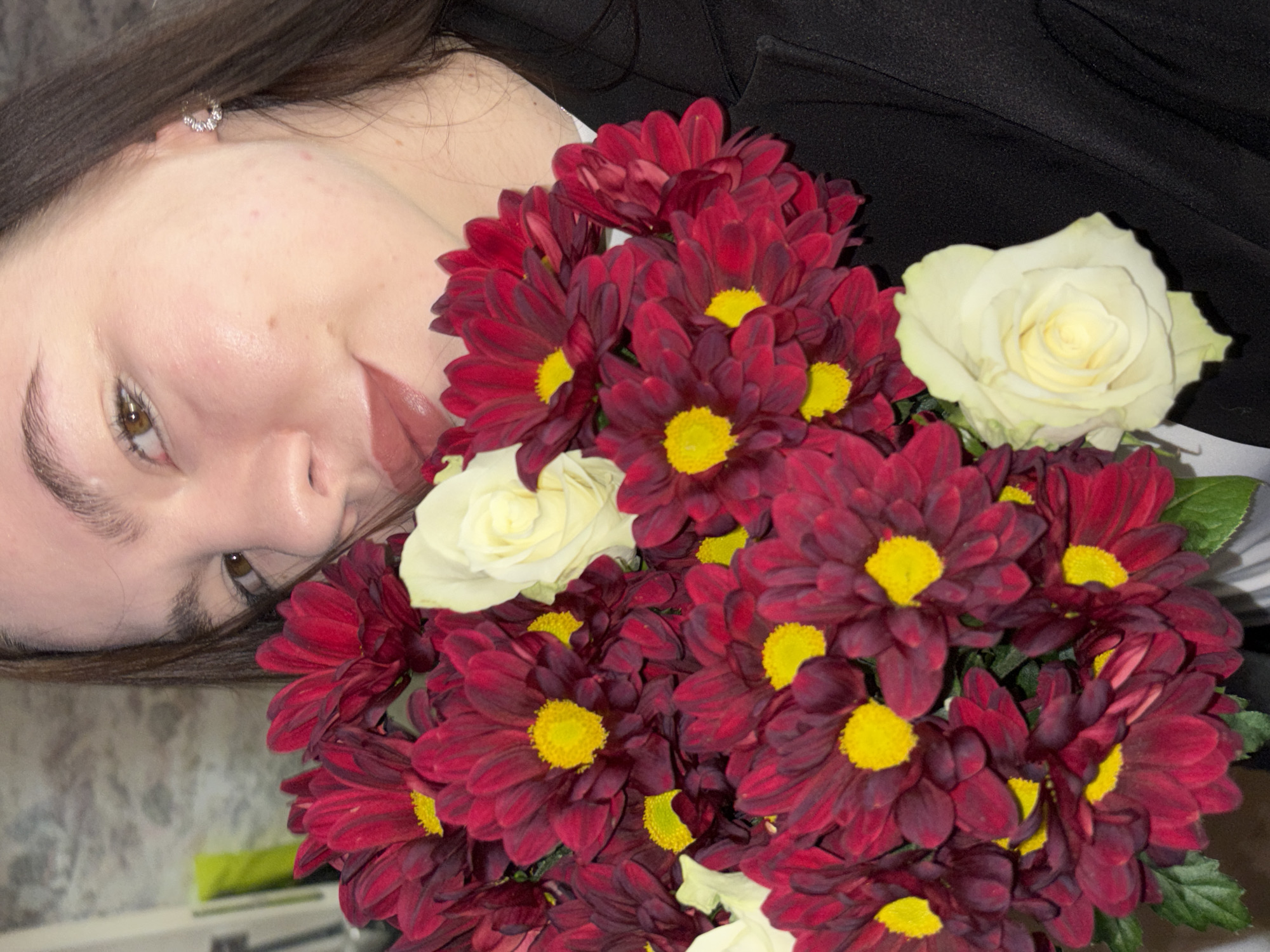 The image shows a woman lying down next to a bouquet of red and white flowers. The woman has light skin, brown hair, and is wearing an earring. The flowers are a mix of red chrysanthemums with yellow centers and white roses. The woman is wearing a black top. The location in the city of Perm, Russia is not evident from the image.