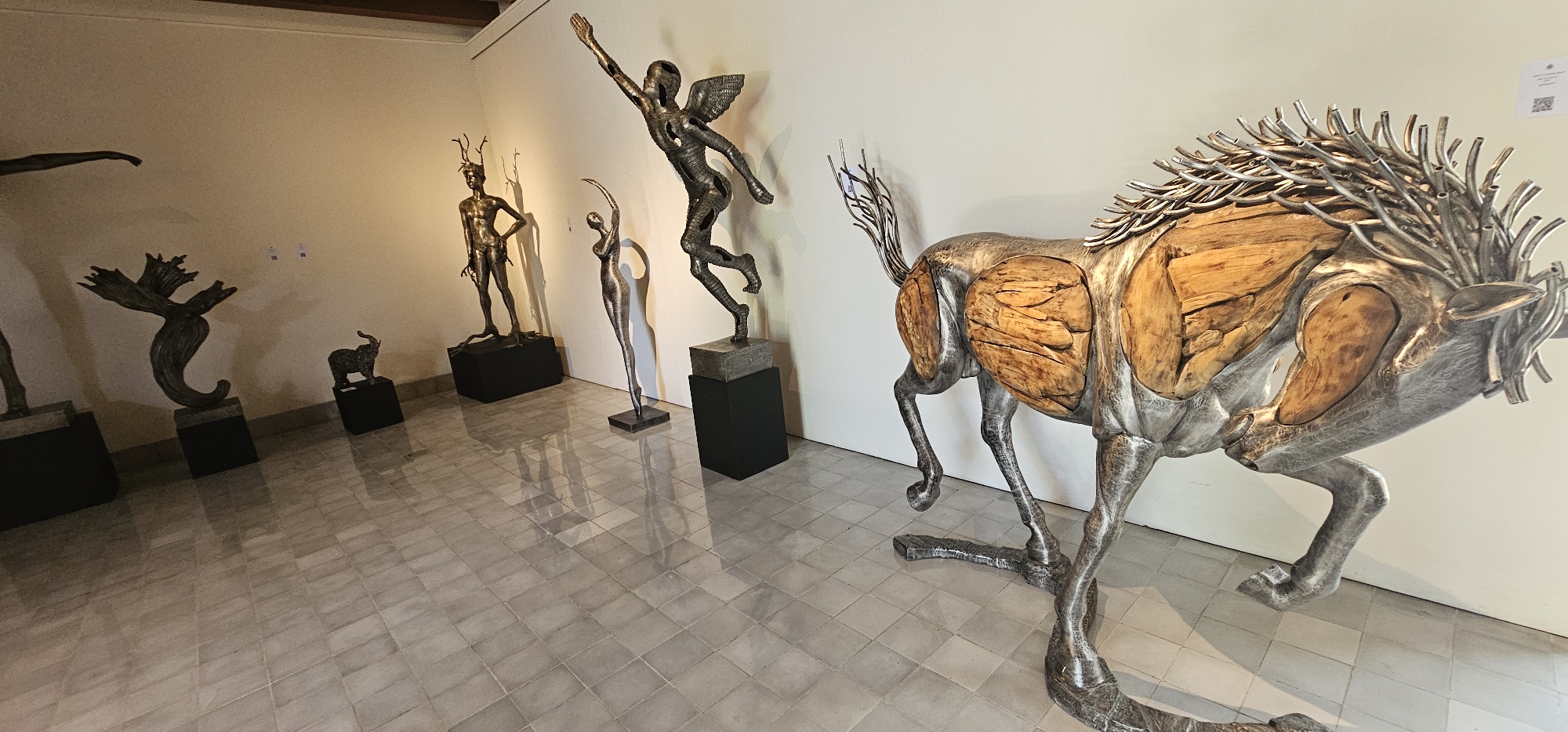 This media file captures an art exhibition in Denpasar, Indonesia, showcasing a collection of contemporary sculptures. The scene is an indoor gallery with light, possibly cream-colored, walls and a reflective, light-toned tiled floor that casts clear reflections of the artworks.
The most prominent subject is a large, dynamic horse-like sculpture positioned on the right in the foreground. It is crafted from mixed materials, featuring a textured, silver-colored metal for its frame and legs, intricately combined with organic, polished wooden inserts that form large sections of its body, suggesting muscle or internal form. Its mane and tail are composed of numerous individual metal rods.
Further into the gallery, along the left wall, several other distinctive sculptures are displayed. These include a metal sculpture of a winged human figure captured in a powerful, upward-reaching pose, standing on a dark pedestal. Adjacent to it, a slender, abstract human form with an arched back and raised arms appears to be dancing or stretching, also made of metal. Another metal sculpture depicts a standing human figure adorned with antlers or horns, placed on a dark pedestal. To its left, a more abstract, swirling, organic metal form is visible, followed by a small, dark animal sculpture, possibly an elephant, on its own pedestal. A partial view of another human-like sculpture can be seen on the far left.
The gallery space is evenly lit, and two small white squares containing QR codes are visible on the wall behind the large horse sculpture. The overall setting is serene, highlighting the intricate details and craftsmanship of the exhibited pieces.
