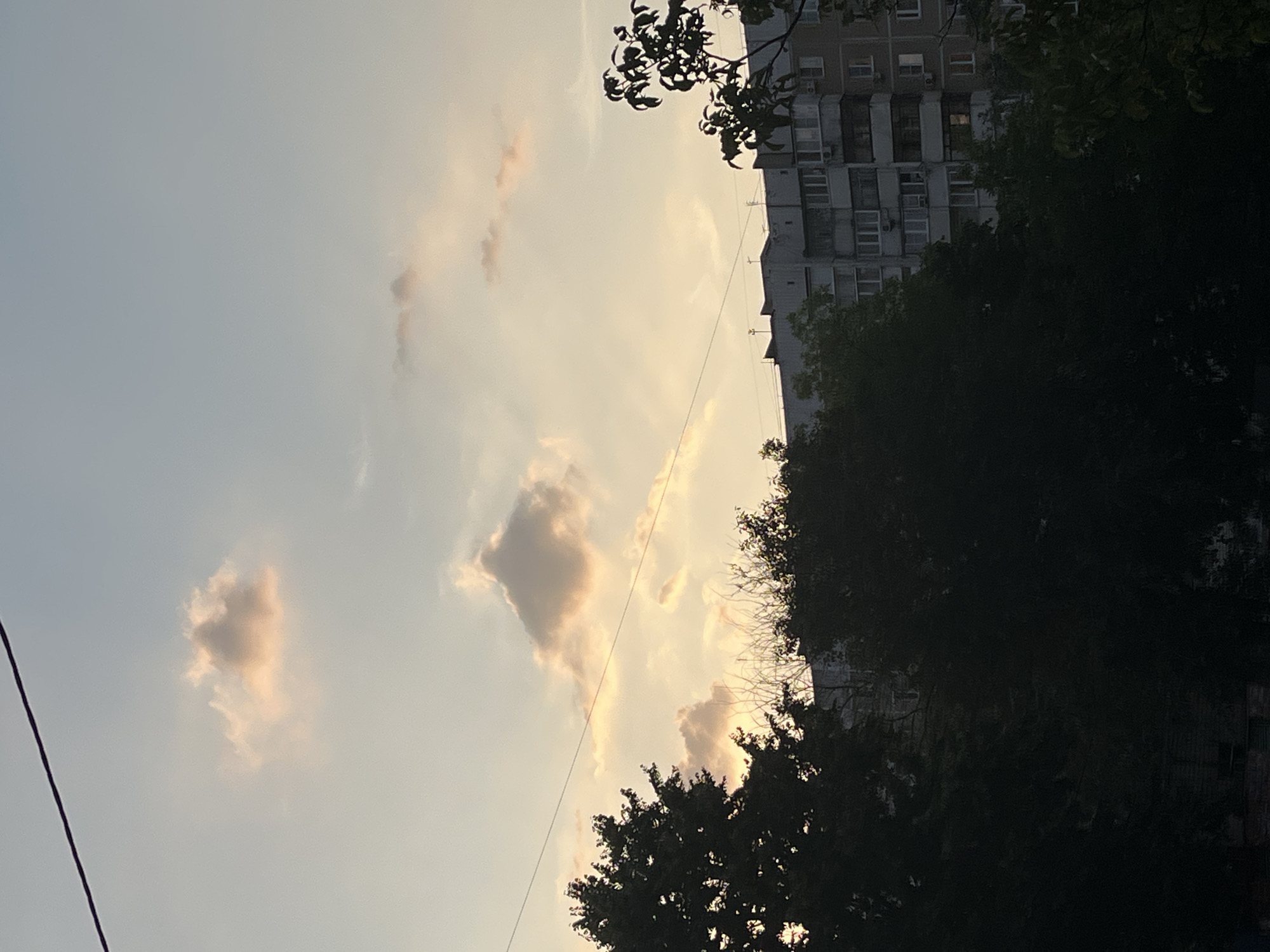 The image depicts a twilight sky over a cityscape. In the sky, several clouds are visible, some tinged with pink and orange hues, suggesting the setting sun. A thin, dark power line stretches diagonally across the upper portion of the frame. Below the sky, silhouetted trees with dark foliage dominate the foreground, partially obscuring the view of a multi-story apartment building in the background. The building appears to be made of concrete and has numerous windows, some of which are illuminated. The overall scene conveys a quiet, urban evening atmosphere.