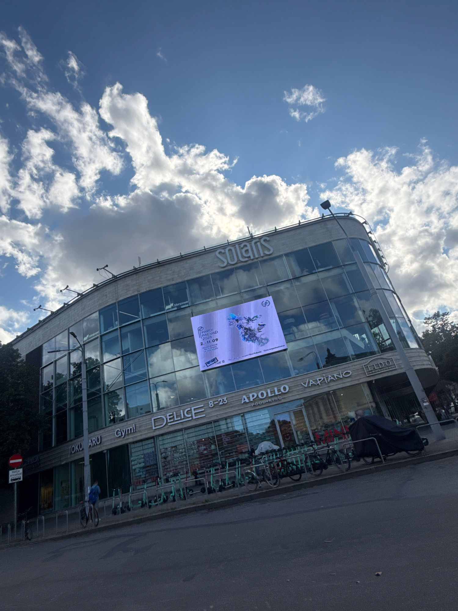 The image shows the Solaris complex located in Tallinn, Estonia, on a cloudy day. The building features a large glass facade with various signs for businesses such as "SOLARIS", "DELICE", "Gym!", "JOKU ARU", "APOLLO FOOD-KINO", and "VAPIANO". A large digital screen is mounted on the building displaying an advertisement. In front of the building, a row of electric scooters and bicycles are parked. A person is seen riding a bicycle on the street. The sky is blue with scattered white clouds, and trees are visible on the left side of the image. A traffic sign is also present on the left.