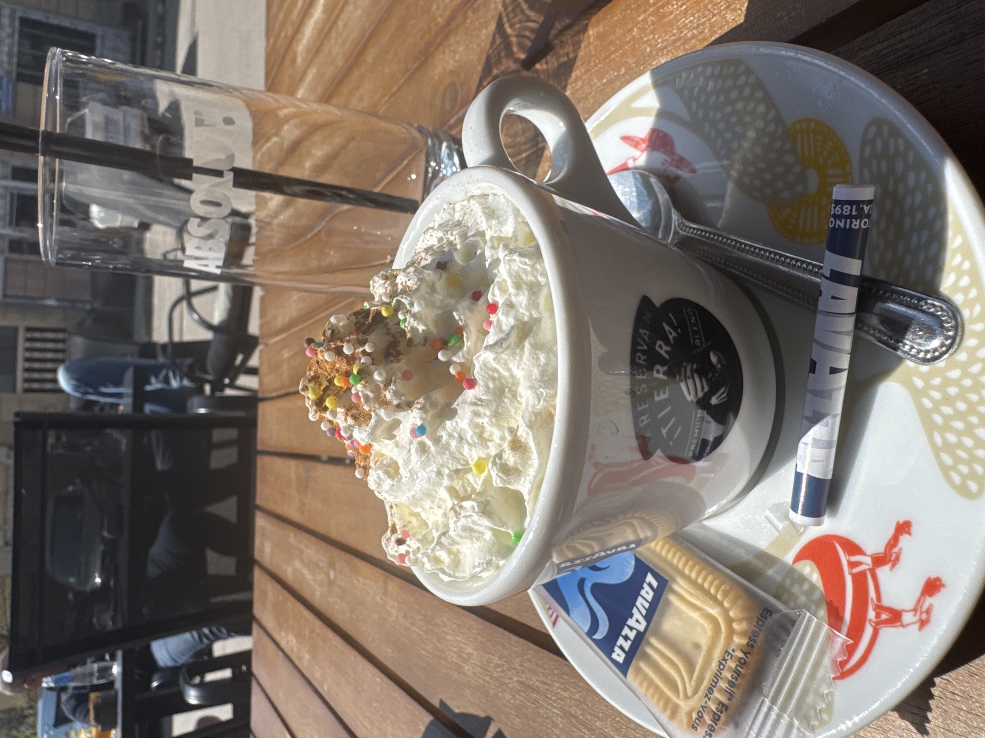 The image shows a table at what appears to be an outdoor cafe or restaurant. On the table, there is a cup of coffee with a large amount of whipped cream and colorful sprinkles, sitting on a saucer decorated with a colorful pattern. A small package of Lavazza biscuits is next to the cup. A spoon rests on the saucer, and a paper sugar packet is also present. To the left of the cup is a tall glass with the word "ABSOLUT" visible on it. The background features an outdoor setting with a glimpse of seating and what looks like a building or fence. The wooden table suggests an outdoor patio or similar setting. There is no information provided in the image that suggests the scene is located in Byala Slatina, Bulgaria.