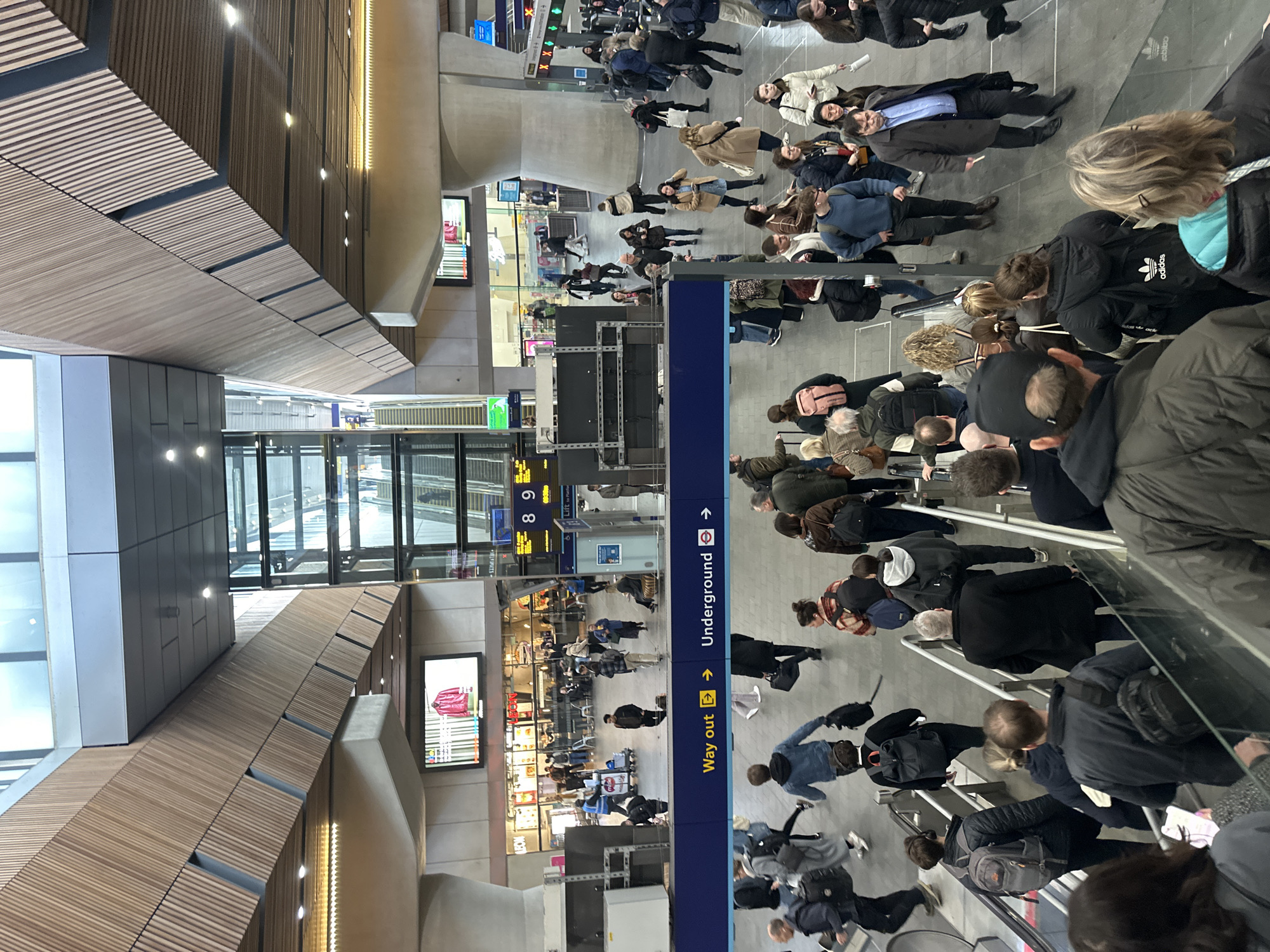 The image portrays a busy, multi-level concourse within London Bridge Station, London, United Kingdom. The viewpoint is elevated, looking down towards lower levels and across the wide interior space.
The environment features modern architecture with high ceilings adorned with light-toned wooden slat paneling on angled and flat surfaces. Recessed lighting fixtures are integrated into the ceiling. Large concrete support pillars and beams are visible throughout. A prominent, tall glass structure, likely an elevator shaft or a bank of escalators, extends vertically through multiple levels in the central background. Digital display screens are present; one shows "8 9" in yellow text, indicative of platform numbers, and another exhibits an advertisement for a red jacket. A dark blue directional sign in the mid-ground clearly indicates "Way out →" and "Underground →" with the distinctive London Underground roundel logo. On a lower concourse level in the background, retail or food establishments are visible, with one displaying the "Wimpy" brand logo.
Numerous individuals, estimated to be over 100, are present across the various levels. They are primarily adults, dressed in diverse casual to smart-casual clothing, including coats, jackets, and sweaters, suitable for urban travel in cool weather. Many carry personal belongings such as backpacks, shoulder bags, and some wheeled luggage. The dominant action is movement, with people walking in multiple directions across the concourse and utilizing escalators, both ascending and descending, in the foreground and background. Some individuals are stationary, possibly waiting or observing. One person in the mid-ground pushes a trolley. No overt personal interactions beyond general navigation are clearly discernible among the crowd.
