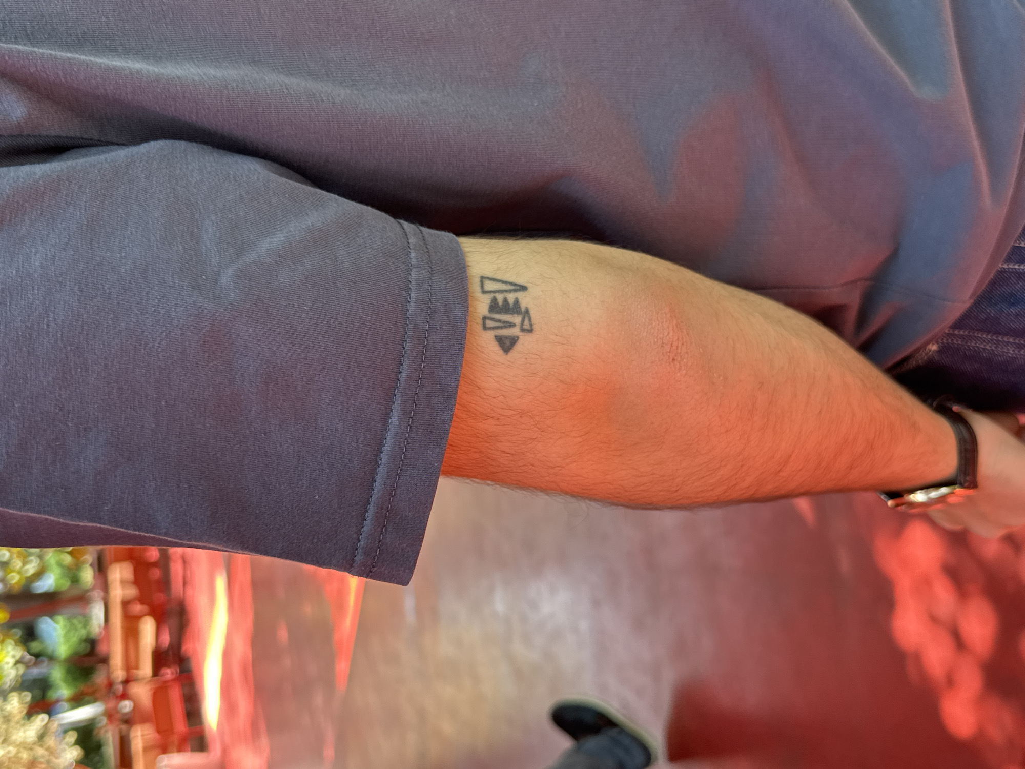 The image shows a close-up of a person's forearm, bearing a small tattoo of abstract geometric shapes. The person is wearing a dark grey t-shirt and blue jeans, and a watch with a dark leather strap is visible on their wrist. The background is blurred, but appears to be an outdoor setting with red-colored flooring and tables and chairs, possibly in a cafe or restaurant. The lighting suggests it may be daytime, with warm, reddish hues dominating the scene. No specific location in Yerevan, Armenia is discernible from the provided image.