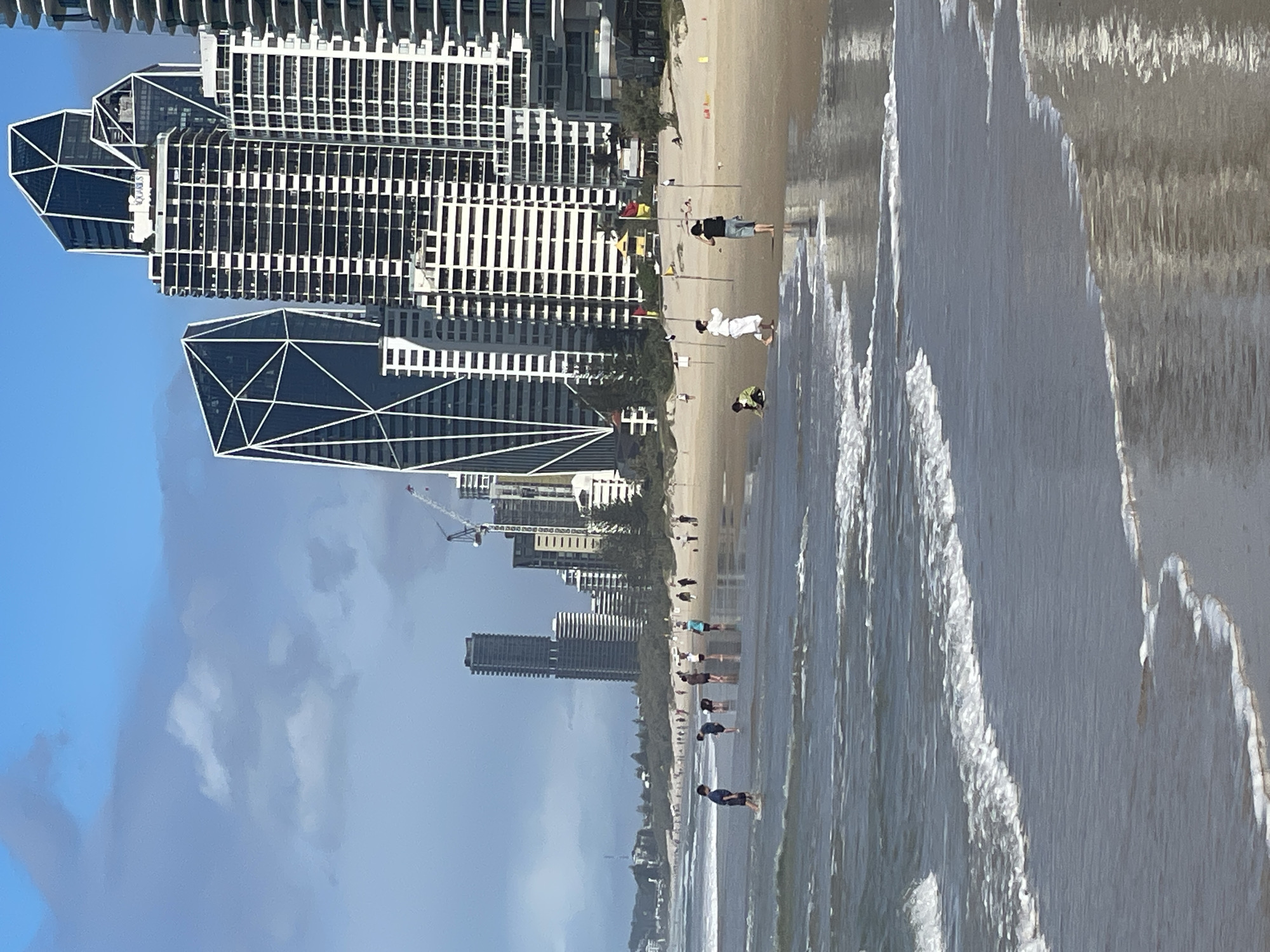 This is a photograph taken at Surfers Paradise, Australia, depicting a beach scene with a backdrop of modern high-rise buildings. The foreground is dominated by the ocean with gentle waves lapping at the sandy shore. Several people are visible on the beach and in the shallow water. In the distance, a cluster of tall buildings, characteristic of the Surfers Paradise skyline, rises against a partly cloudy blue sky. Construction cranes are also visible amongst the buildings, suggesting ongoing development. On the beach, some individuals appear to be walking or standing, while others are interacting in smaller groups. The overall impression is of a typical day at a popular urban beach.
