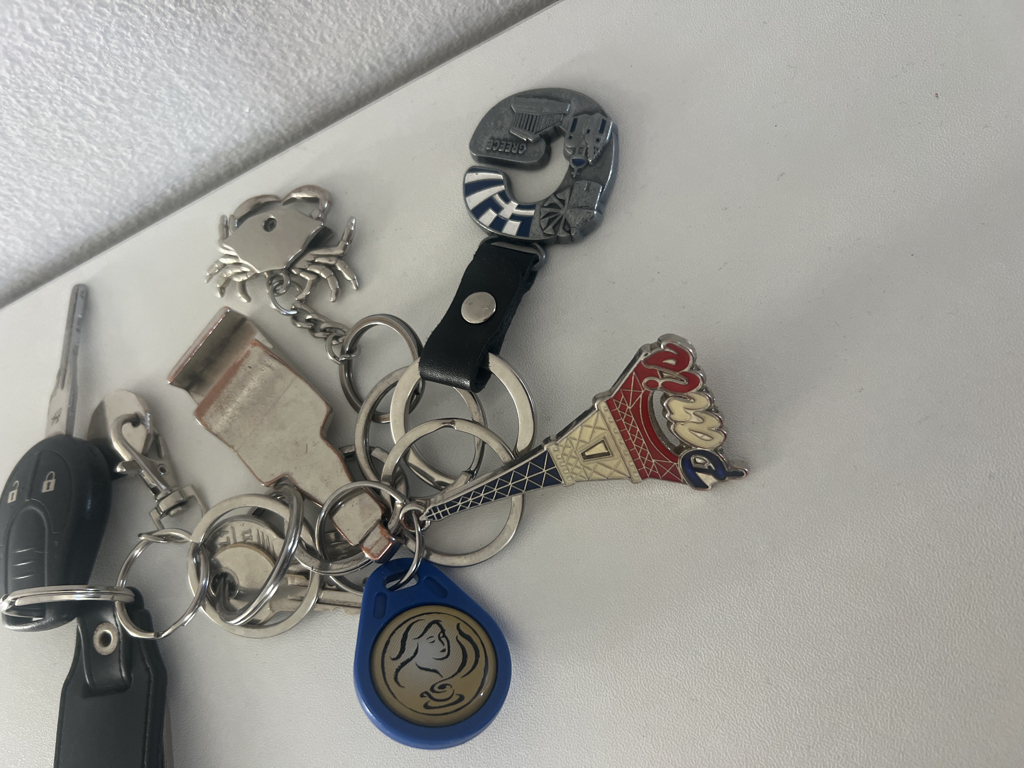 A close-up, brightly lit, still-life image shows a set of keys and keychains lying on a white surface. The items appear to be suspended or lying on a countertop or similar flat surface. The keys appear to be an automotive key and a standard house key. The keychains consist of the following designs: a crab, a red white and blue tower shape, a blue tag containing the image of a woman, and a piece in the shape of the letter C with “Greece” labeled. Gorleston-on-Sea, United Kingdom is not portrayed in the image.