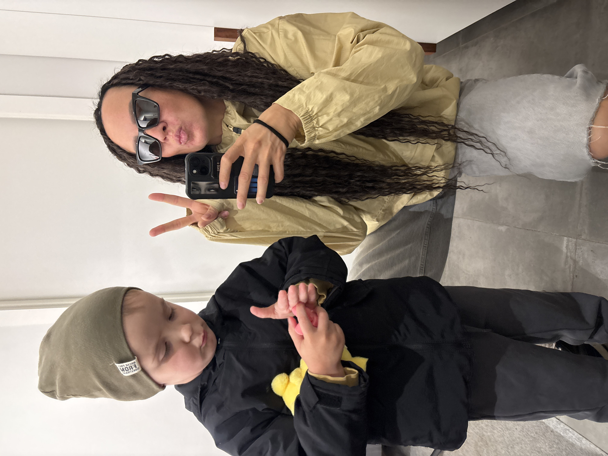 This vertical mirror selfie, captured in Chaikovsky, Russia, features an adult woman and a young child indoors. The woman, positioned at the top of the frame, has long, dark, curly hair and wears black rectangular sunglasses. She is dressed in a light yellow or beige hooded jacket and light grey distressed jeans. She holds a black smartphone in her right hand, taking the picture, and flashes a peace sign with her left hand while playfully pursing her lips.
Below her, a child, likely a toddler, is seen from the chest up. The child wears a dark olive green beanie with a white tag, a black zip-up jacket, and dark pants. They are intently looking down at their hands, which are holding a small pink object. A fluffy yellow object, possibly a toy, is visible near their right wrist.
The background consists of plain white walls and a grey tiled floor, suggesting a clean, modern indoor environment. The image is brightly lit, indicating good indoor lighting. The visible text on the child's beanie tag, when unmirrored, appears to read "BORN FREE FROM GORGEOUS". The overall mood is casual and playful, capturing a candid moment between what seems to be a parent and child.