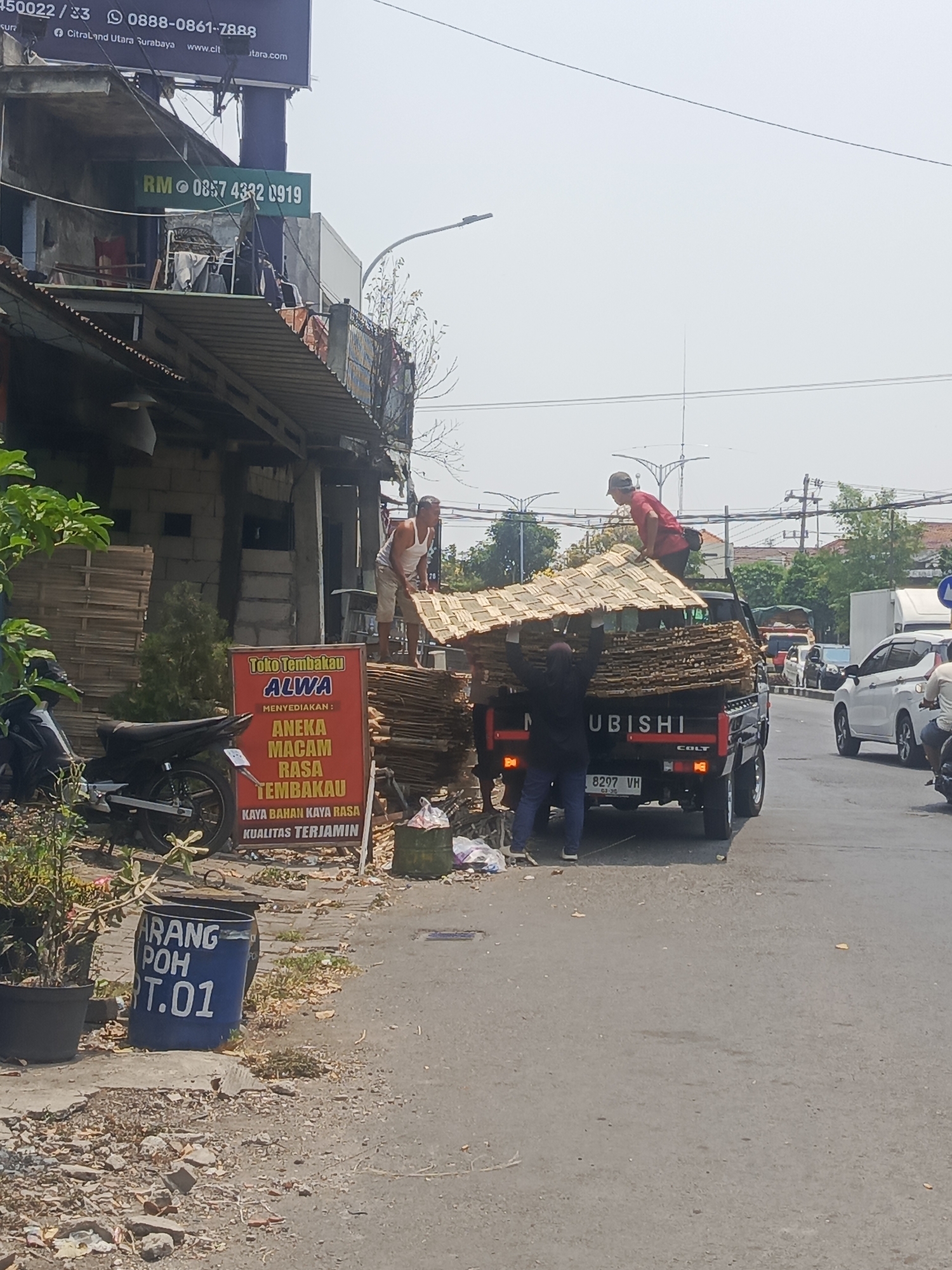 The image depicts a street scene in Surabaya, Indonesia, likely during daylight hours. The foreground features a blue barrel with white lettering that reads "ARANG POH T.01", indicating it may be related to charcoal. To the left, a motorcycle is parked near a potted plant. A red and white sign with Indonesian text, "Toko Tembakau ALWA MENYEDIAKAN: ANEKA MACAM RASA TEMBAKAU KAYA BAHAN KAYA RASA KUALITAS TERJAMIN", is visible, suggesting a tobacco shop.
In the middle ground, a black Mitsubishi Colt pickup truck is loaded with what appears to be bundled bamboo or reeds, possibly roofing material. Three individuals are actively involved in loading or unloading these materials. One man in a white tank top stands on a pile of the material, while two others, one in a red shirt and another in dark clothing, are near the truck bed, assisting with the load.
The background reveals a street with other vehicles, including a white car and other trucks, indicating moderate traffic. Overhead, power lines and streetlights are visible, along with some buildings and trees. The overall atmosphere suggests a commercial area with ongoing activity. The billboard in the upper left corner contains text that includes "450022/33 0888-0861-7888" and "Citraland Utara Surabaya www.clt.com", which helps to confirm the location as Surabaya.