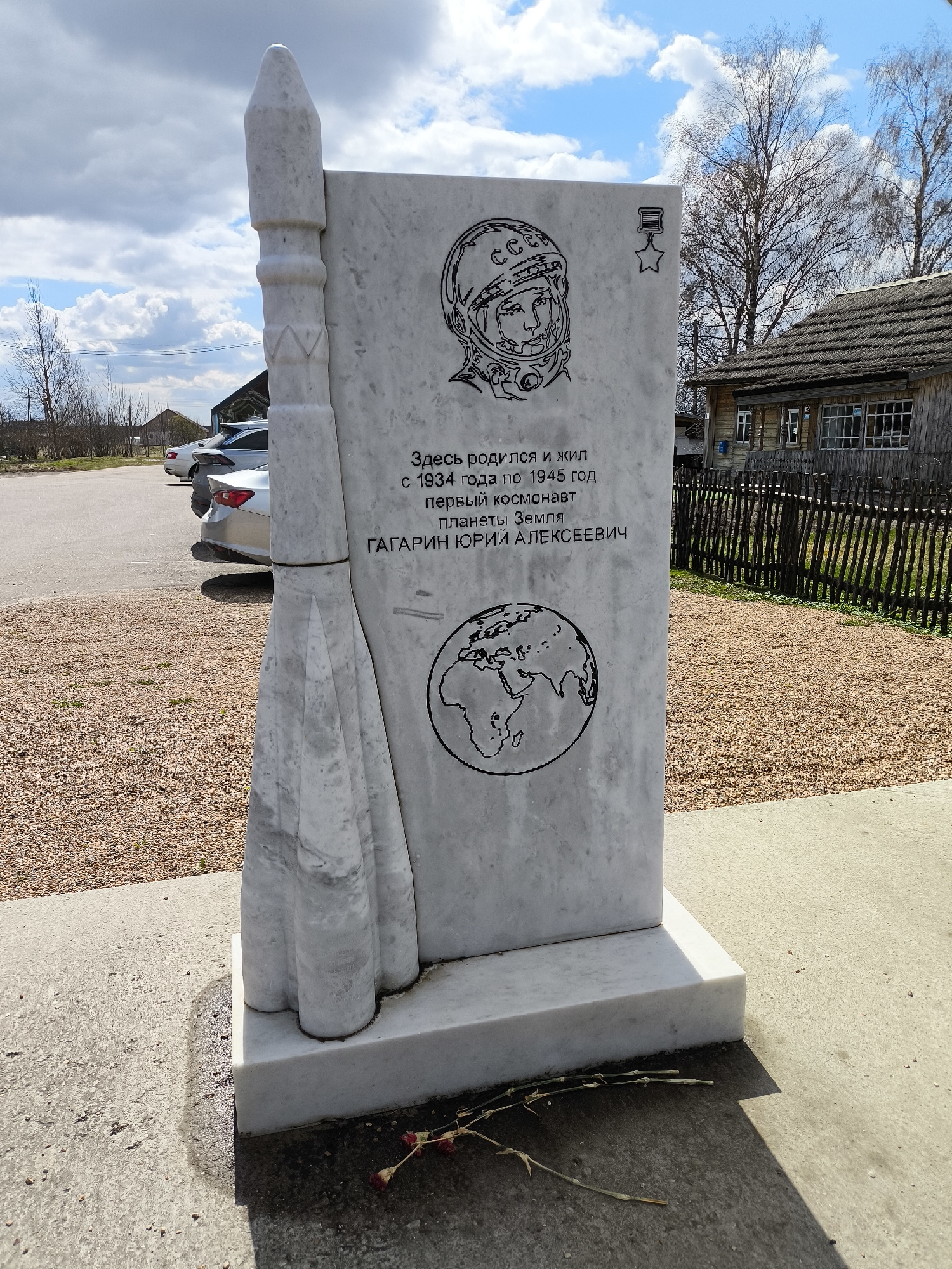 This image captures a memorial monument dedicated to Yuri Gagarin in Kluschino, Russia, under a bright, partly cloudy sky. The monument is made of light-colored marble, featuring a tall, rectangular slab with a stylized rocket carved vertically along its left edge, resting on a broader base.
Etched onto the main slab are several designs and Cyrillic text. At the top, there is a detailed outline of a cosmonaut's helmet, with "CCCР" (USSR) faintly visible on it, and to its right, an emblem of a medal with a star. Below these, the inscription reads: "Here was born and lived / from 1934 to 1945 / the first cosmonaut / of planet Earth / GAGARIN YURIY ALEKSEEVICH." Below the text, an outline of the globe, depicting Earth's continents, is etched.
The setting appears rural or semi-rural. In the background to the right, a traditional wooden house with a thatched roof and a wooden fence is visible. To the left, several modern cars are parked on a gravel area. Bare trees stand against the sky, suggesting early spring. The ground immediately around the monument consists of a concrete path, with some withered red flowers or foliage placed at its base. The scene is illuminated by strong daylight, casting shadows from the monument onto the ground.
