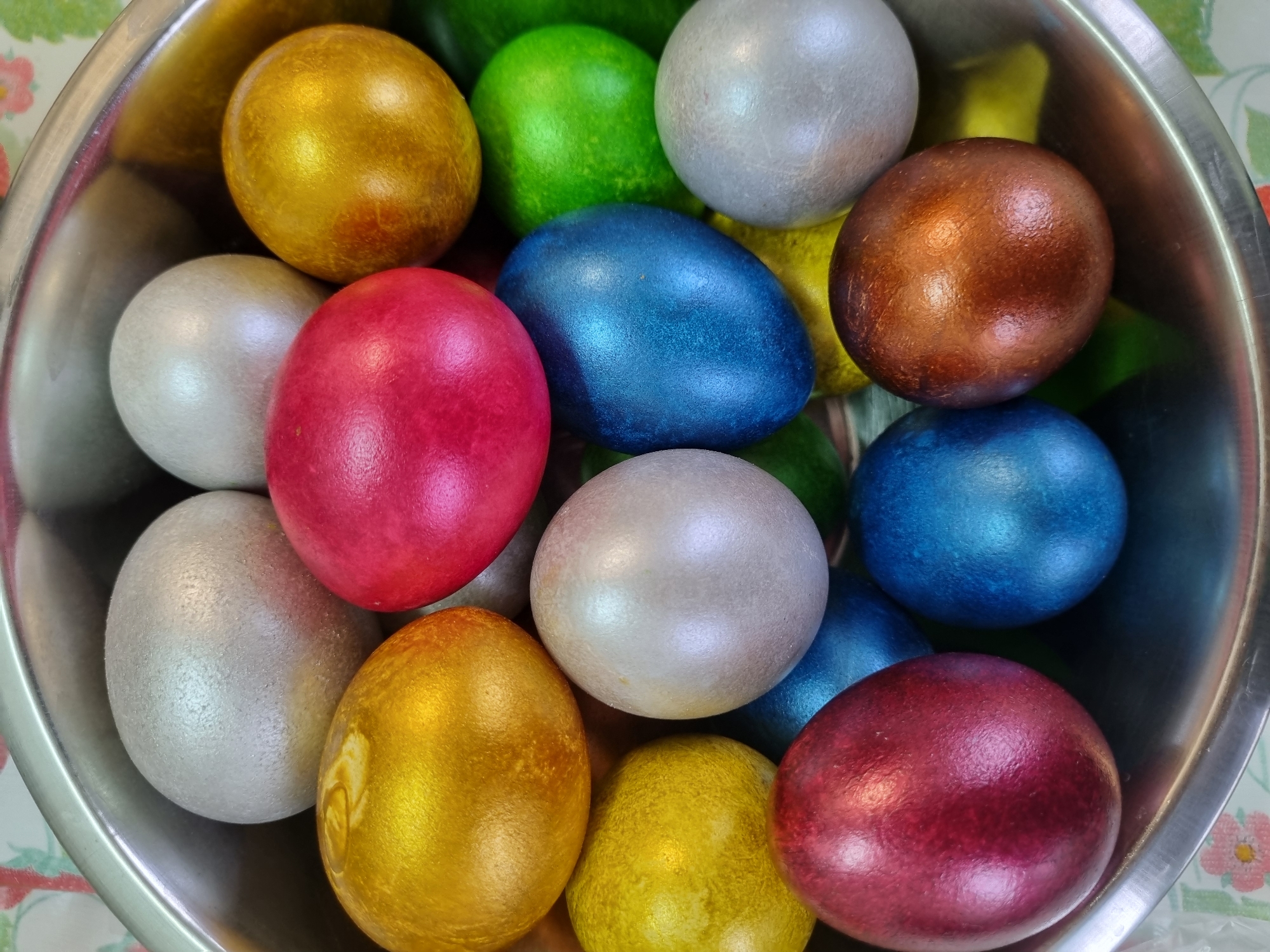 A high-angle, close-up shot depicts a collection of dyed Easter eggs in a metal bowl. The eggs are various colors including gold, green, silver, red, blue, and bronze. The metallic sheen suggests they may have been dyed with a pearlizing or metallic finish. The bowl is situated on a surface covered with a light-colored floral pattern cloth. The image does not depict a location in Kemul, Russia, or any other identifiable location or people.