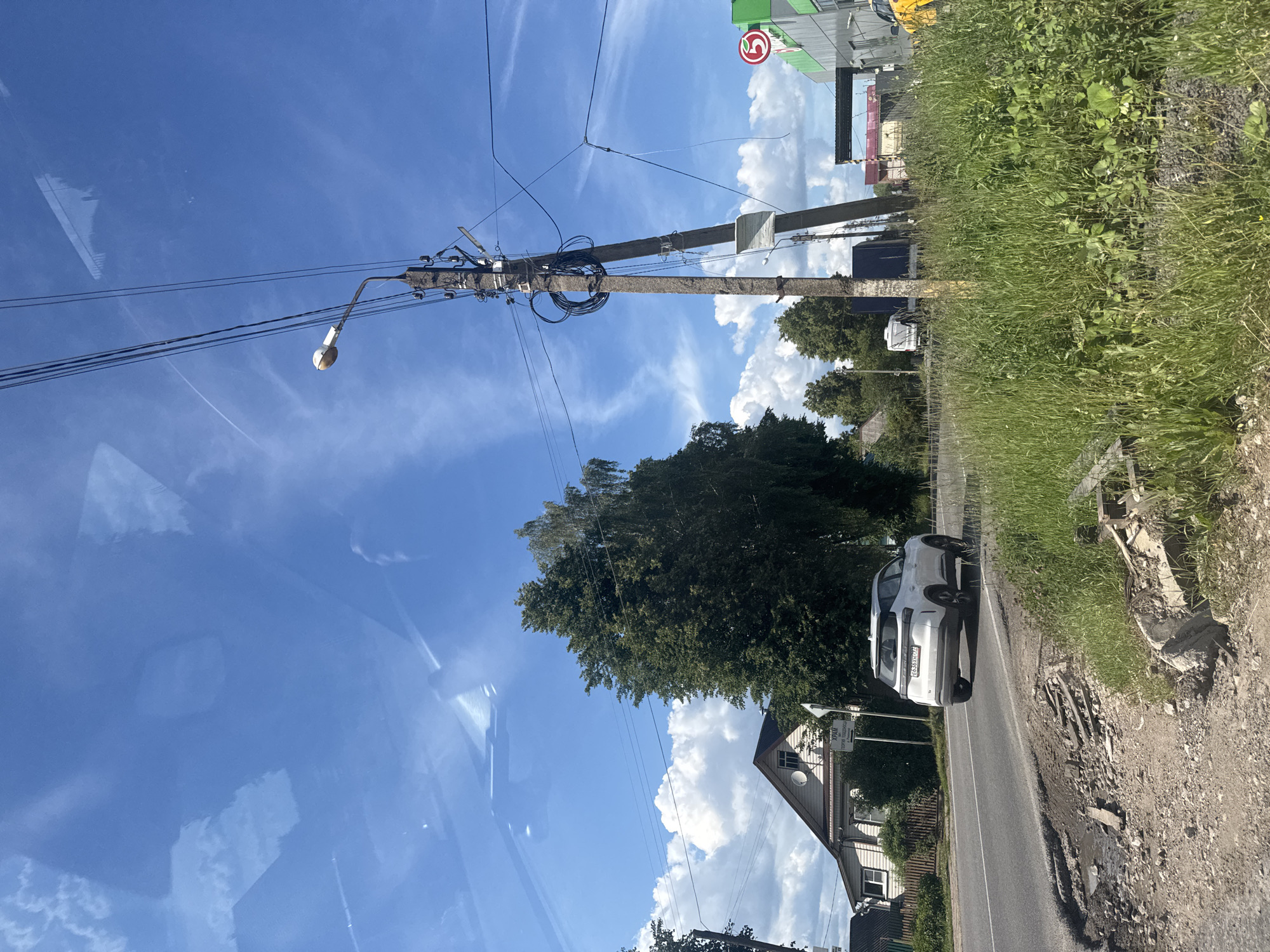 The image depicts a street scene in Medyn, Russia, on a clear day with a blue sky and scattered clouds. A white SUV is driving on a paved road that shows signs of disrepair, with a pothole and debris along its edge. To the left of the road, a light-colored house with a gable roof is visible, surrounded by trees and a fence. In the foreground on the right, tall green grass and vegetation grow, with a concrete utility pole and overhead power lines dominating the center of the frame. A streetlamp is attached to the pole. In the background, several other vehicles, including a white van and a dark truck, are seen on the road. A building with green and white signage, possibly a store, is situated further down the street. A red circular sign with a white number "5" is affixed to the building. The overall environment suggests a rural or semi-urban setting with a mix of residential and commercial elements.