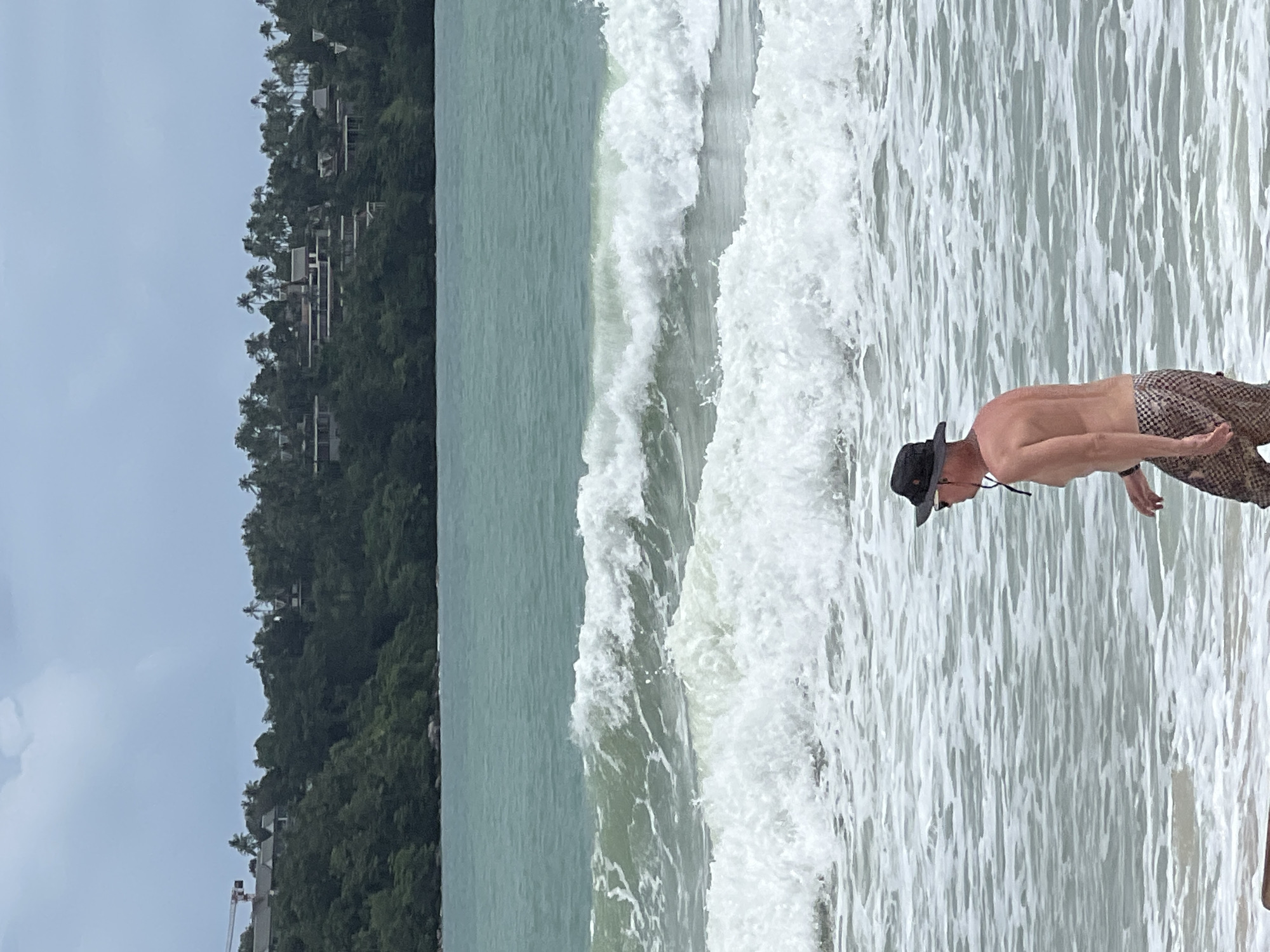The image shows a beach scene. A man wearing a hat and patterned pants is standing in the surf. The ocean waves are crashing near him. In the background, there is a heavily forested hillside with some buildings visible. A construction crane is seen on the left side of the image. The sky is overcast. The location is Unknown, Unknown.