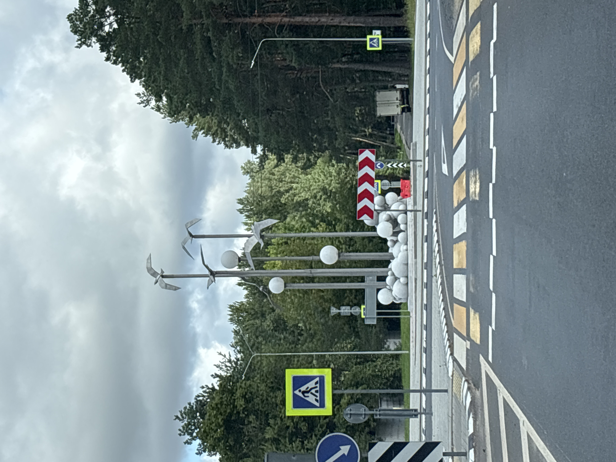 The image depicts an outdoor scene with a focus on a roundabout and a sculpture. In the foreground, a multi-lane asphalt road with clear pedestrian crossing markings is visible. The roundabout itself is delineated by white and black striped curbs.
Dominating the center of the image is a sculpture composed of multiple metallic bird figures in flight, mounted on poles of varying heights. Spherical elements, likely decorative, are clustered at the base of the sculpture and also appear on some of the poles. The sculpture is situated within the roundabout.
Surrounding the roundabout and extending into the background are numerous trees, primarily coniferous with some deciduous foliage, suggesting a natural or park-like setting. A street lamp is visible on the right side of the image.
Several traffic signs are present: a blue circular sign with a white arrow pointing left is on the left side, and a yellow square sign with a blue pedestrian symbol is also on the left. On the right side, a similar yellow square pedestrian sign is visible. Additionally, a red and white chevron directional sign is mounted on a pole to the right of the sculpture, indicating a change in road direction.
A light-colored truck is parked on a road in the background, further into the trees. The sky above is overcast with gray clouds. The overall environment suggests a public area, possibly a town square or a park entrance, within the Ждановіцкі сельскі Савет, Belarus. There are no visible people actively engaged in any interactions or actions within the frame.
