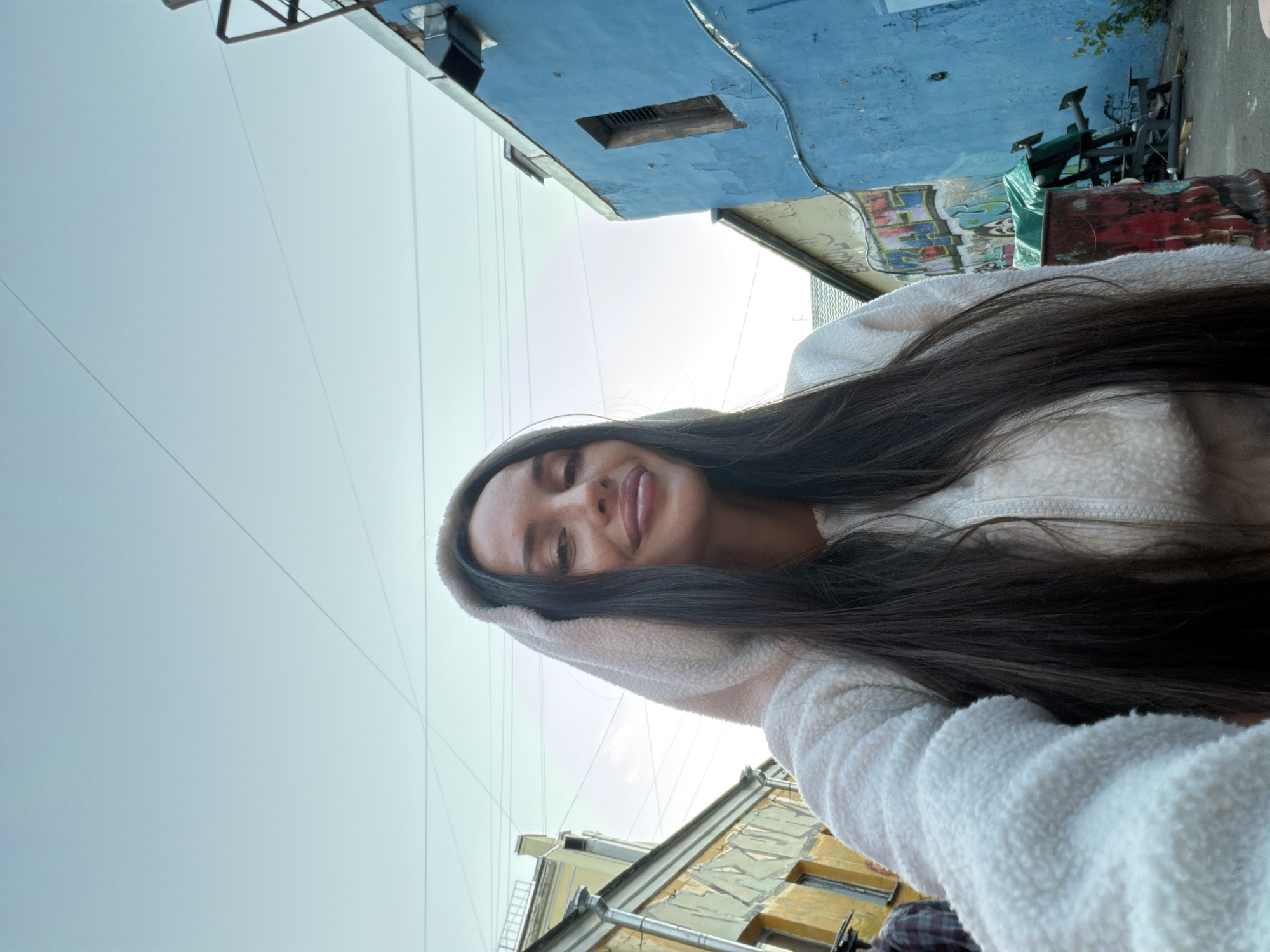 A low-angle, full-body selfie taken outdoors in a narrow urban alleyway in Saint Petersburg, Russia, features a young woman with long dark hair wearing a cream-colored fuzzy hoodie. The woman is smiling and looking directly at the camera, her head tilted slightly. She is positioned in the foreground, with her right arm and shoulder visible. The background consists of two buildings facing each other across the alley. The building on the left is yellow and shows signs of wear and tear, with peeling paint and graffiti. The building on the right is painted a vibrant blue and has a rustic, weathered appearance, also with visible graffiti. The sky above is overcast and pale blue. Various objects are visible in the alley, including a red barrel with graffiti, a green tarp covering some equipment, and some electrical wires strung across the sky. The overall impression is an informal, candid snapshot of a person in a somewhat gritty urban environment.