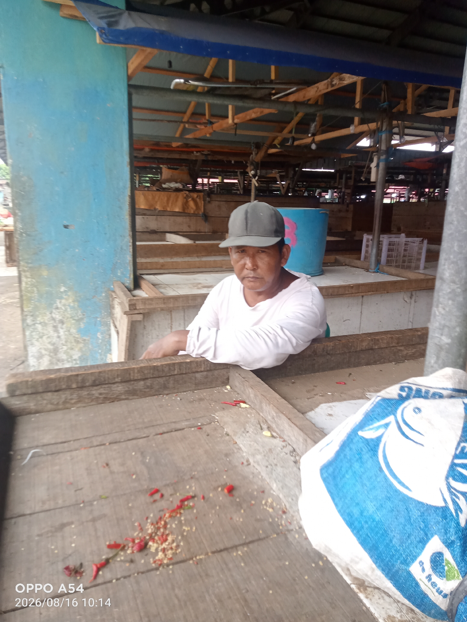 The image depicts a street market stall in Jambi, Indonesia, likely on the morning of August 16, 2026. A person wearing a grey cap and a light-colored long-sleeved shirt is seated behind a wooden stall. Red chili peppers and seeds are scattered across the wooden surface of the stall. A large blue and white woven bag with a fish graphic is also visible in the foreground. The background shows the market structure with wooden beams and a blue tarp. The date and time stamp "OPPO A54 2026/08/16 10:14" is superimposed on the bottom left corner of the image.