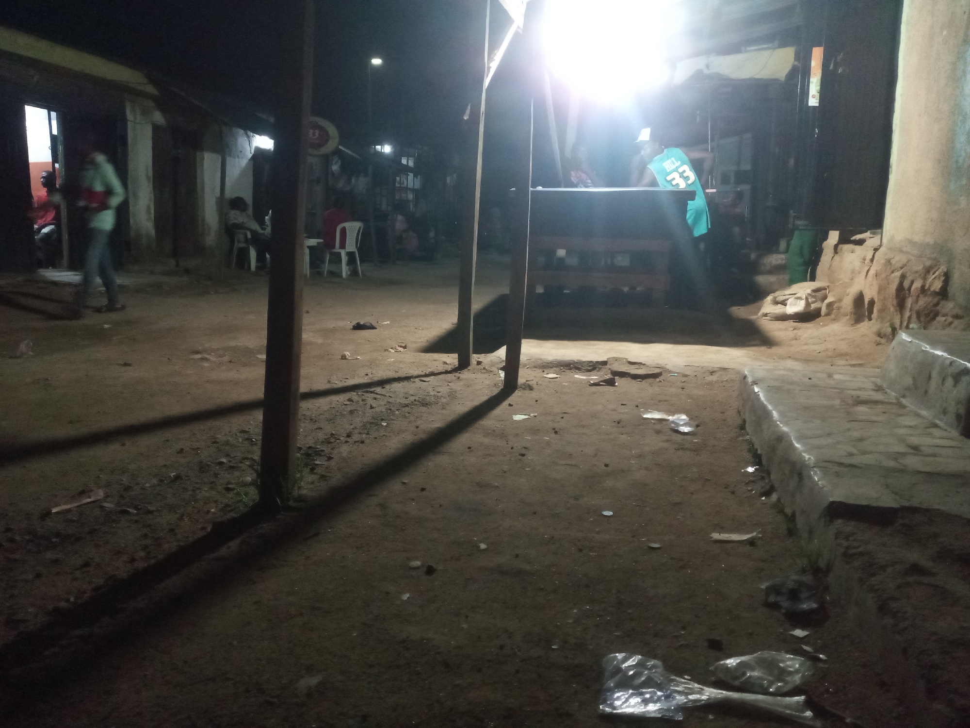 The image depicts a dimly lit outdoor space in Jos, Nigeria, likely at night. The ground is uneven, dirt, littered with debris including plastic bags and small objects. Light is provided by a bright overhead light source, casting long shadows from wooden support posts. In the background, blurred figures are visible, seated and standing near buildings, including one wearing a turquoise jersey with the number "33." The buildings appear to be simple, possibly shops or residences. There is a counter or table with a flat surface behind the posts. The scene suggests a public area with a blend of commercial and residential activity. A concrete step-like structure runs along the right edge of the image. The overall atmosphere is one of informal gathering and activity.
