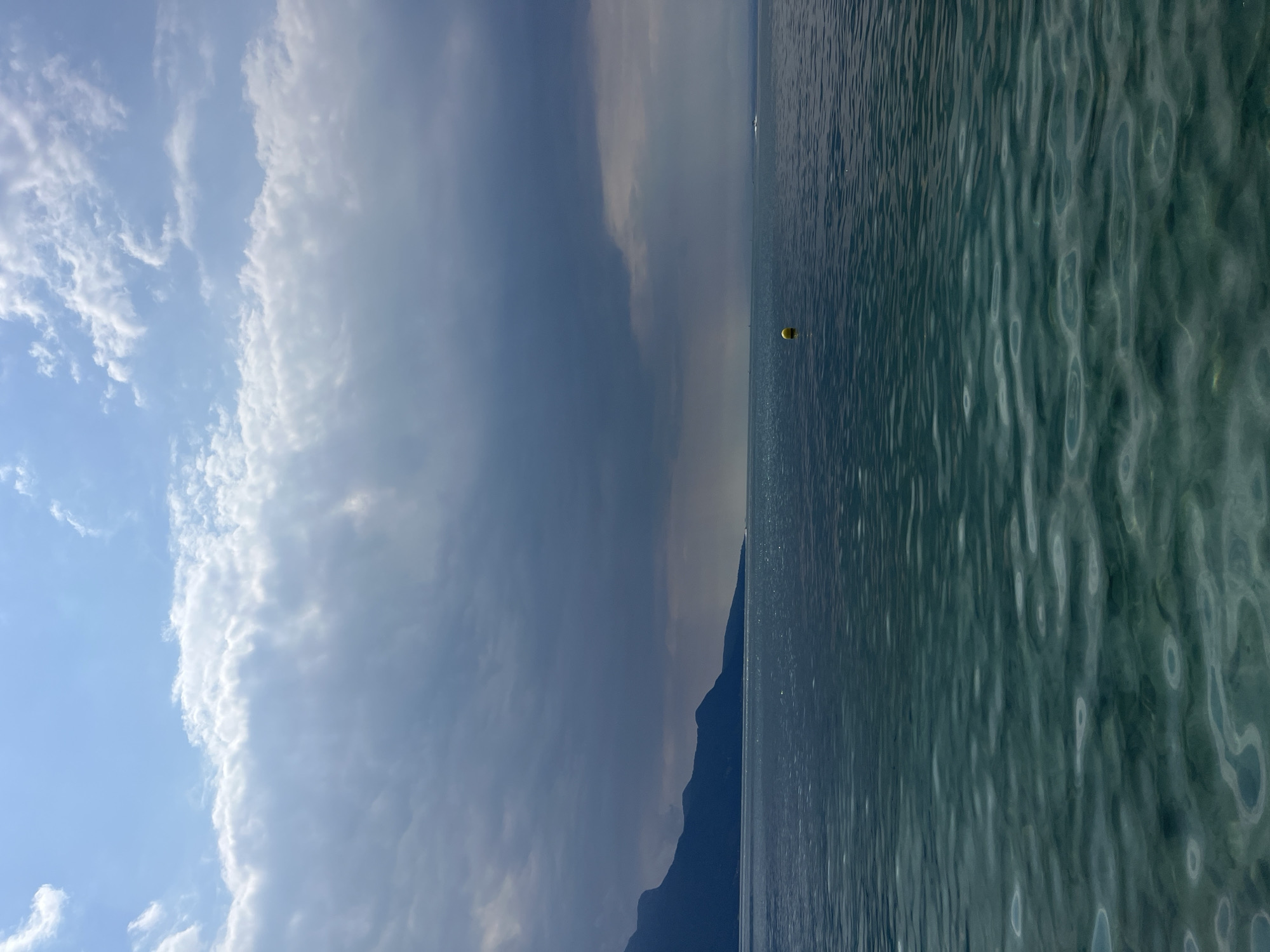 This image depicts a body of water, likely a lake, with a mountainous landscape on the left side. The sky is partly cloudy, with dramatic dark clouds in the center and lighter clouds towards the top. In the middle of the water, a yellow buoy is visible. The water itself appears calm with ripples on the surface. The distant shoreline on the left features dark, silhouetted mountains. On the right side of the frame, there is a suggestion of movement in the water, possibly a boat wake, and a distant shoreline. The overall impression is a serene, natural landscape. Given the context provided, this scene is located in La Tour-de-Peilz, Switzerland, a town situated on the shores of Lake Geneva, which is bordered by the Alps.