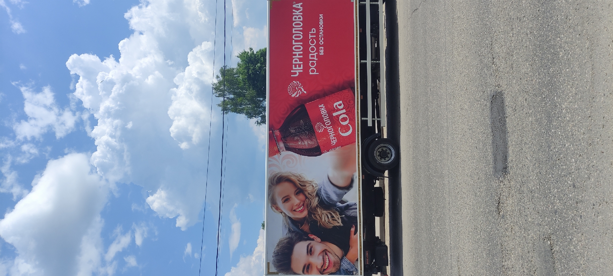 On a sunny day with a partly cloudy sky, a billboard advertising "Chernogolovka Cola" is displayed on the side of a white truck. The billboard features a young, smiling couple taking a selfie, with a bottle of cola and the brand's logo prominently displayed. The truck is parked on an asphalt road in Fryazino, Russia, with visible potholes in the foreground. Trees and power lines are visible in the background.