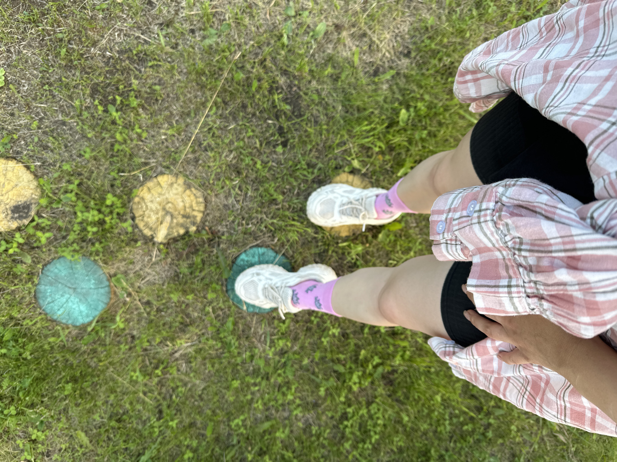 A top-down shot shows a person's legs in white sneakers and pink socks standing on grass. In the background, there are three painted wooden discs on the grass, one teal and two yellow. The person is wearing a black skirt and a pink and white plaid shirt. The scene appears to be outdoors, possibly in a park or garden. The location is specified as Yaroslavka, Russia.