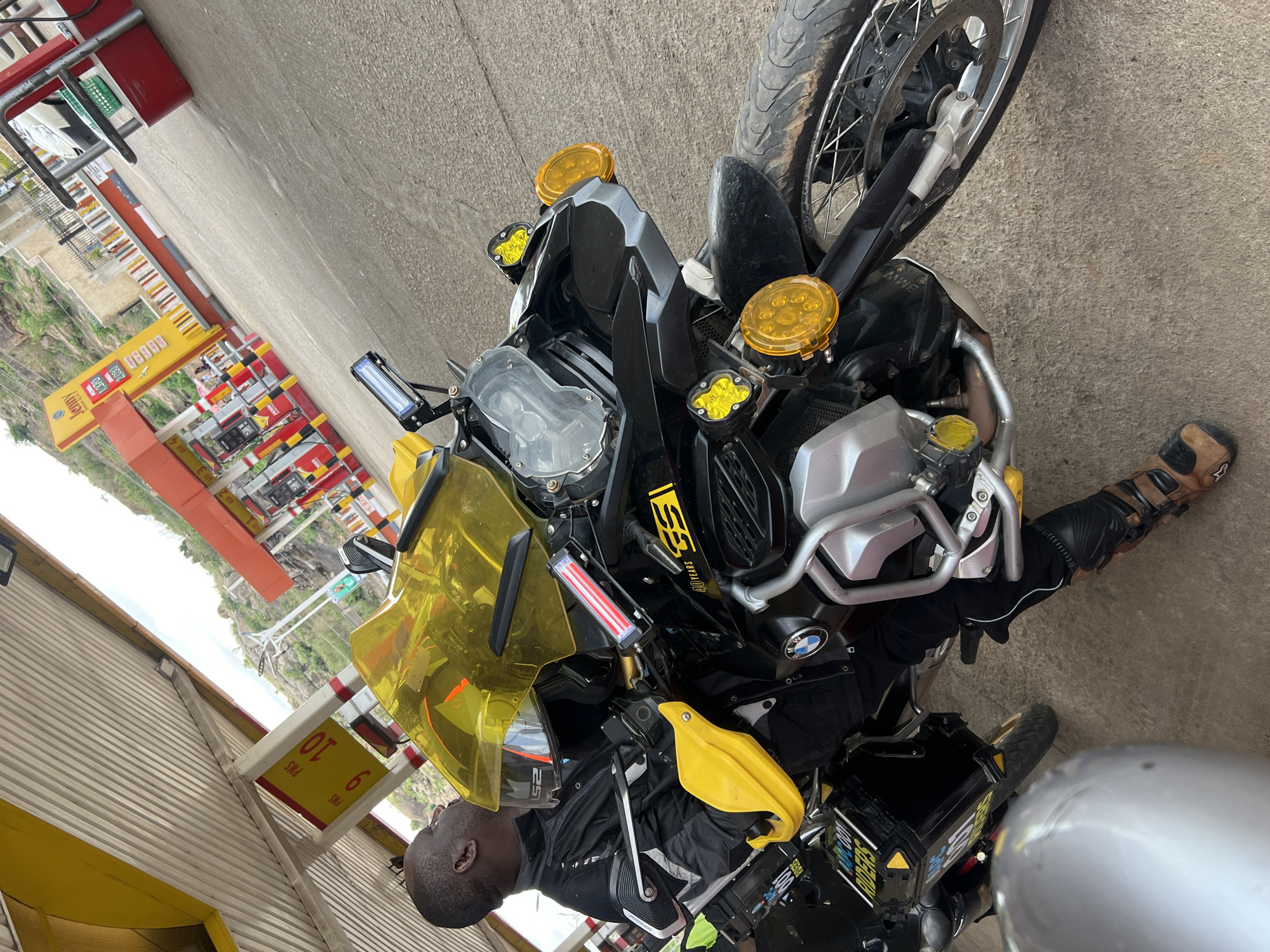 The image depicts a man standing next to a black and yellow BMW motorcycle at a Jenney gas station in Gura Top, Nigeria. The man is wearing a black jacket, black gloves, and brown motorcycle boots. The motorcycle has additional lights and accessories attached. The gas station has several fuel pumps and a sign displaying the price of fuel. The surrounding environment includes buildings and vegetation.