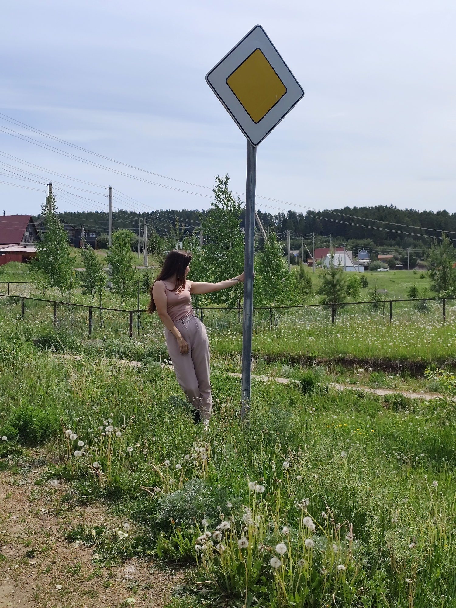 A woman in a tan tank top and tan pants leans against a yellow and white diamond-shaped signpost in a grassy field. The location is Фоки, Russia. The field is filled with dandelions. A dirt path is visible in the lower left corner. Behind the field is a fence, trees, houses, and telephone poles. The sky is overcast.
