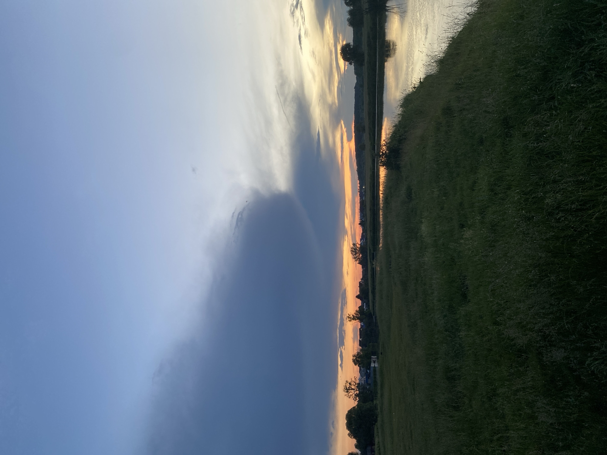 The image depicts a serene landscape scene at dusk in the city of Tujmazy, Russia. The sky is a gradient of blues and oranges, with dramatic dark clouds on the left transitioning to lighter, wispy clouds on the right, illuminated by the setting sun. A calm body of water, likely a river or lake, occupies the right side of the frame, reflecting the colors of the sky. The left and middle sections of the image feature a grassy field with tall, uncut grass, suggesting a rural or undeveloped area within or near the city. In the distance, a line of trees and the rooftops of several houses are visible, indicating the presence of a settlement. The overall atmosphere is peaceful and natural, with no people or animals present in the frame. The lighting suggests it is either sunrise or sunset.