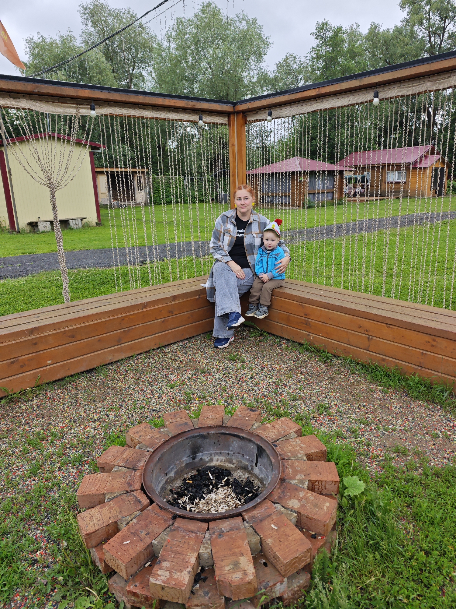 This image depicts an outdoor recreational area in Malyi Bukor, Russia, under an overcast sky. In the foreground, a circular fire pit is constructed from reddish-brown bricks arranged around a metal basin, which contains ash and burnt white debris. The ground around the fire pit consists of multicolored gravel mixed with patches of green grass and weeds.
In the midground, a woman and a young child are seated on an L-shaped wooden bench, constructed from multiple horizontal dark-stained planks. The adult female, with reddish hair pulled back, wears a light-colored plaid shirt over a dark t-shirt, light blue jeans, and dark athletic shoes with white soles and pink accents. Her left arm is positioned around the child. The child, appearing to be a toddler, wears a blue zip-up jacket, brown pants, gray and blue athletic shoes, and a distinctive hat with red, white, and yellow colors, featuring a pom-pom. The child's right arm is around the woman's waist. Both individuals are looking directly towards the camera.
Above the bench, a wooden beam structure supports vertical strands of light-colored material, resembling ropes or chains, forming a decorative screen. Two exposed white lightbulbs are suspended from this beam. To the far left in the midground, beyond the bench, a light yellow building with a red trim is visible, alongside a decorative, tree-like structure made of twisted material. A small, simple wooden bench rests near the base of this building. Further back on the right, two wooden cabins with dark red roofs are situated on a grassy lawn. One cabin features a visible porch or deck. Lush green deciduous trees form a dense backdrop across the entire upper portion of the image, extending to the horizon under a pale gray, cloudy sky. An orange flag is partially visible in the upper left corner.