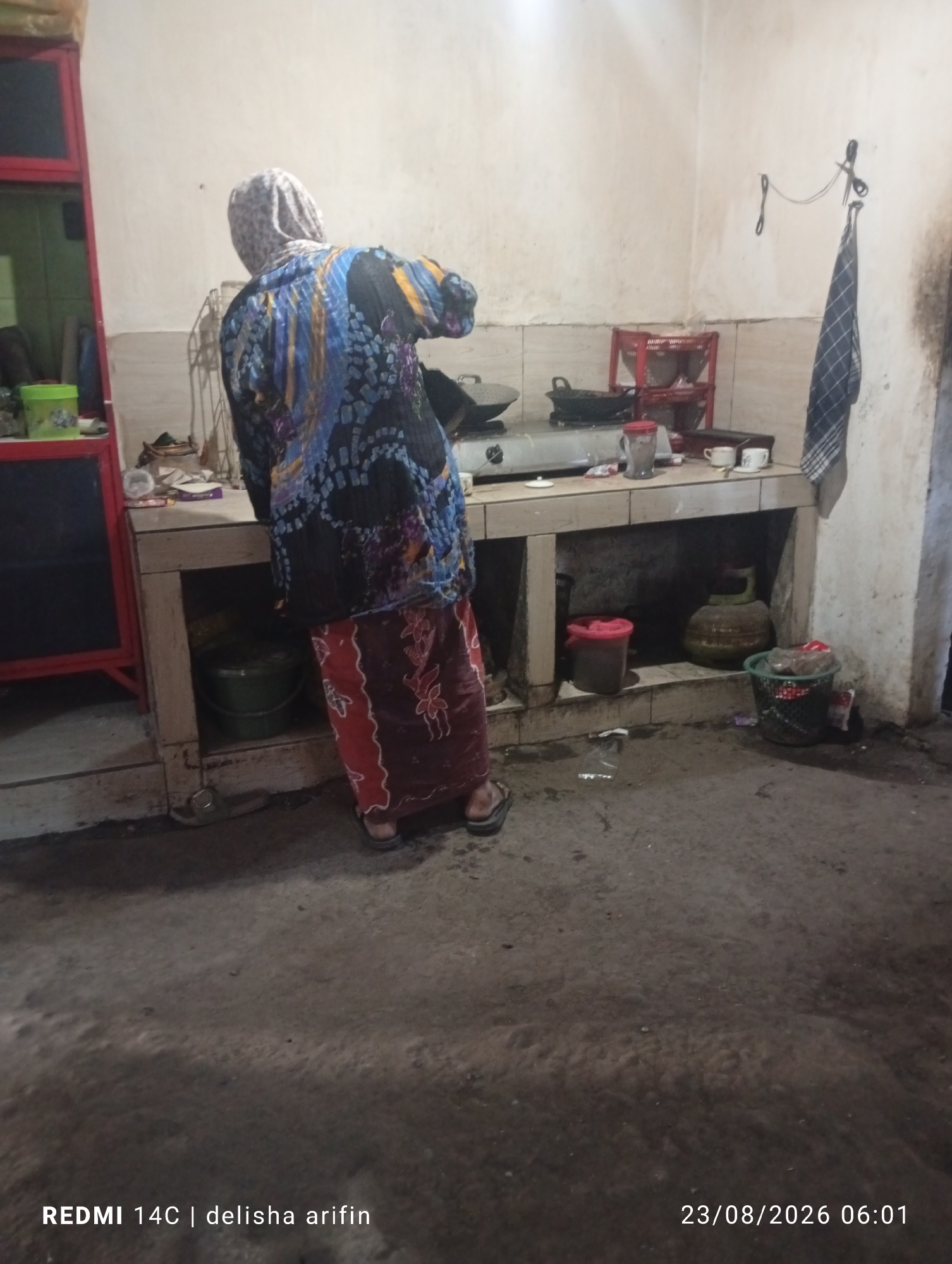 The image depicts an indoor scene, likely a kitchen, in Tamansari, Indonesia, on August 23, 2026, at 06:01. A person, viewed from behind, is standing at a counter, possibly engaged in cooking or food preparation. The individual is wearing a patterned headscarf and a long, colorful tunic over a red and white patterned skirt. The kitchen counter is made of what appears to be concrete or tiled material and has a stovetop with two burners. Various items are placed on the counter, including pots, a red-lidded container, cups, and a container with a grey lid. Below the counter, there are storage areas containing a green bucket, a red bucket, a green gas cylinder, and a green bin with some red and white items inside. To the left, a red-framed shelving unit is partially visible, with a green container on one of its shelves. The floor appears to be concrete and shows signs of dirt and debris. A blue and white checkered cloth hangs on the wall to the right, suspended by a string attached to a hook, with a pair of scissors also attached to the wall nearby. The overall environment suggests a utilitarian and somewhat cluttered domestic space.