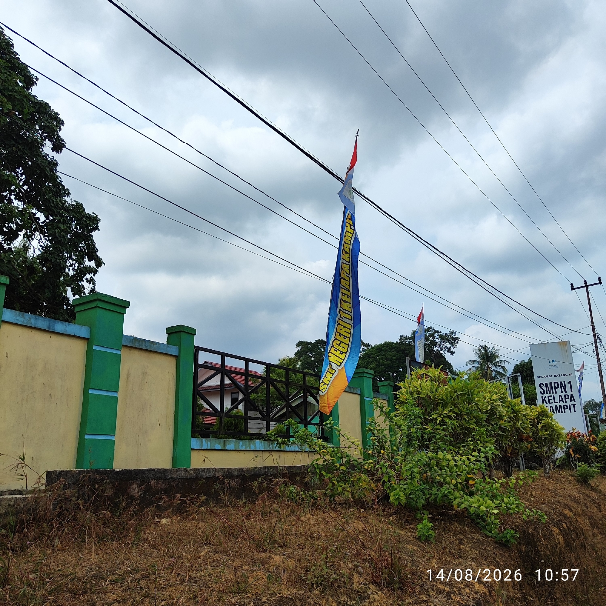 An outdoor, low-angle, medium shot captures a cloudy day in Unknown, Indonesia, with a white sign reading "SELAMAT DATANG DI SMPN 1 KELAPA KAMPIT" on the right. A tall, green-and-yellow fence with blue trim runs horizontally across the frame, bordering a dark-colored fence with a white building behind it. Several power lines crisscross the sky. A blue and yellow flag with red and white elements hangs from a pole, slightly to the left of the center. Dense green foliage is visible on the right side of the image, and dry grass and dirt occupy the foreground. A dark tree with green leaves is on the left. The timestamp "14/08/2026 10:57" is visible in the lower right corner. No people are present.