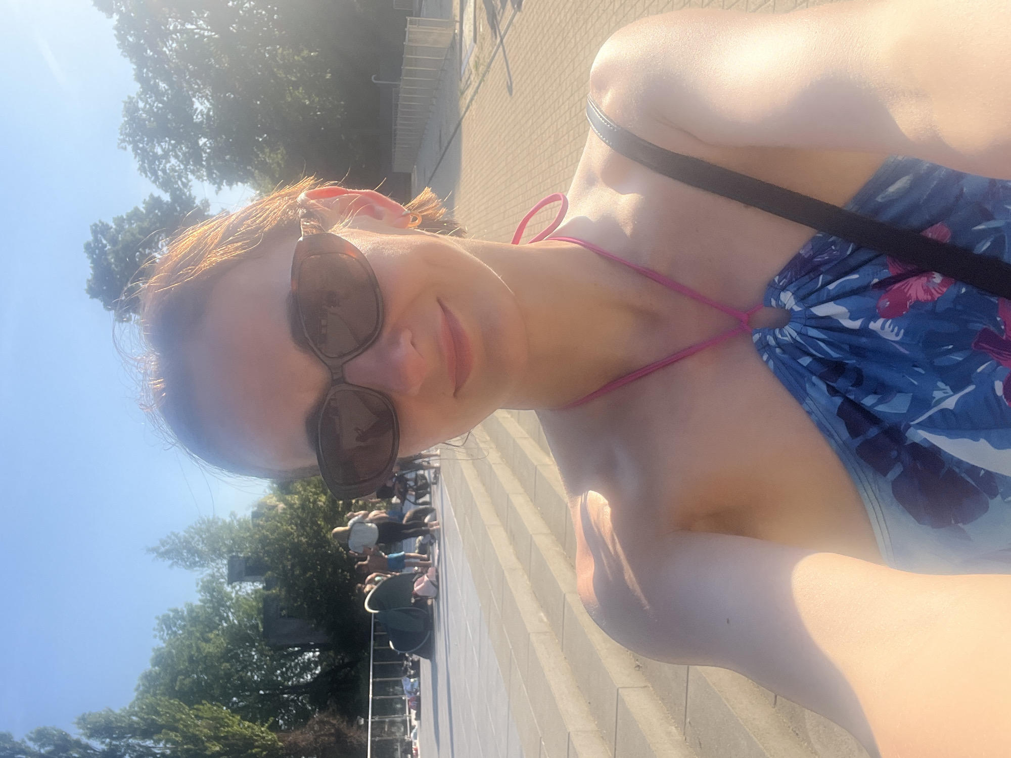 A woman wearing sunglasses and a blue floral swimsuit takes a selfie in an outdoor setting. She is smiling and looking at the camera. The background shows a plaza with steps, trees, and other people. The sun is shining brightly, casting a lens flare across the image. The location is not specified.